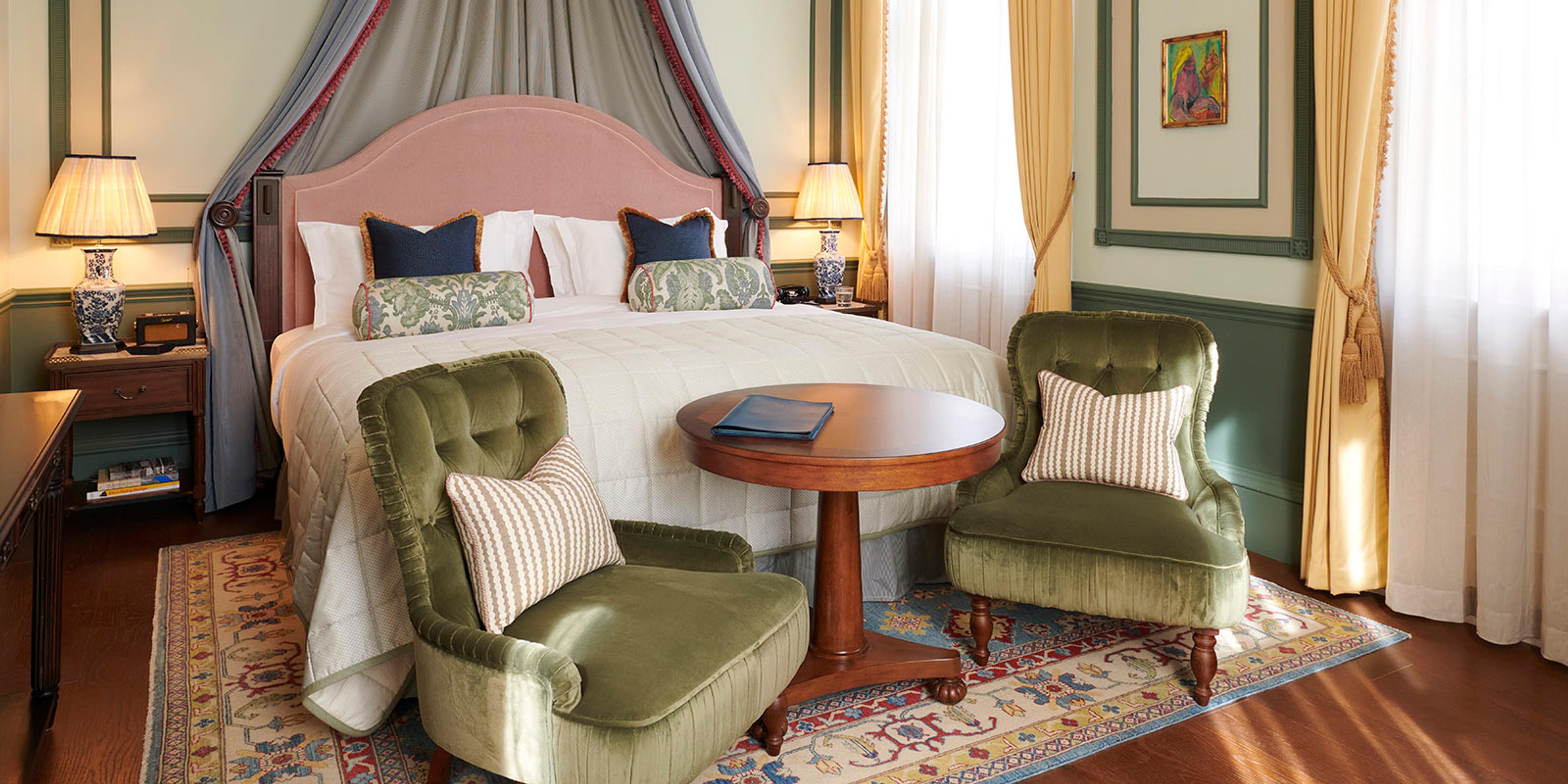 a canopied bed with a pink headboard and grey drapes, flanked by green armchairs around a round table on a patterned rug