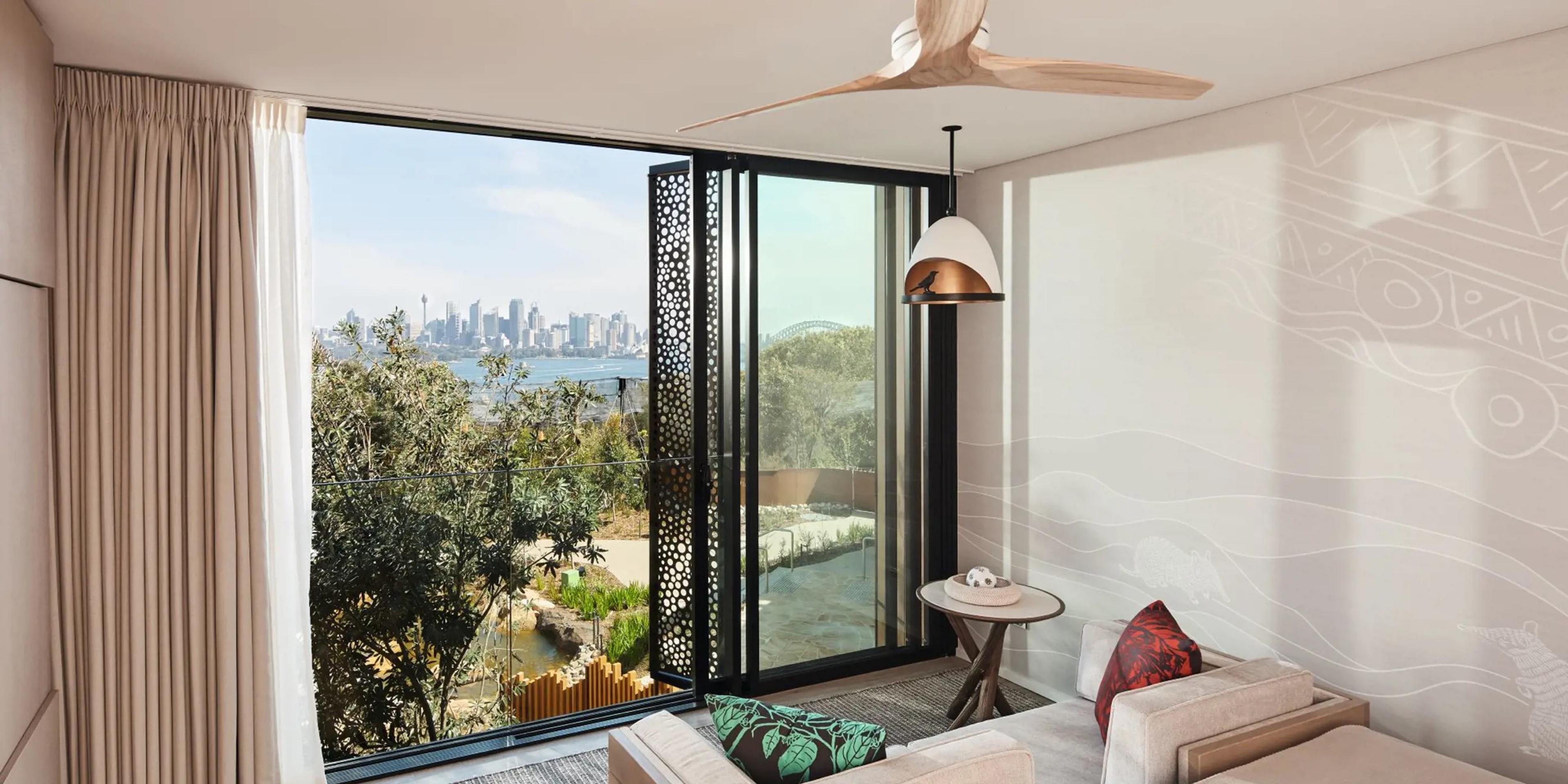 A bedroom sofa beneath glass doors folded open to a balcony, with the Sydney Harbour Bridge visible above the trees.