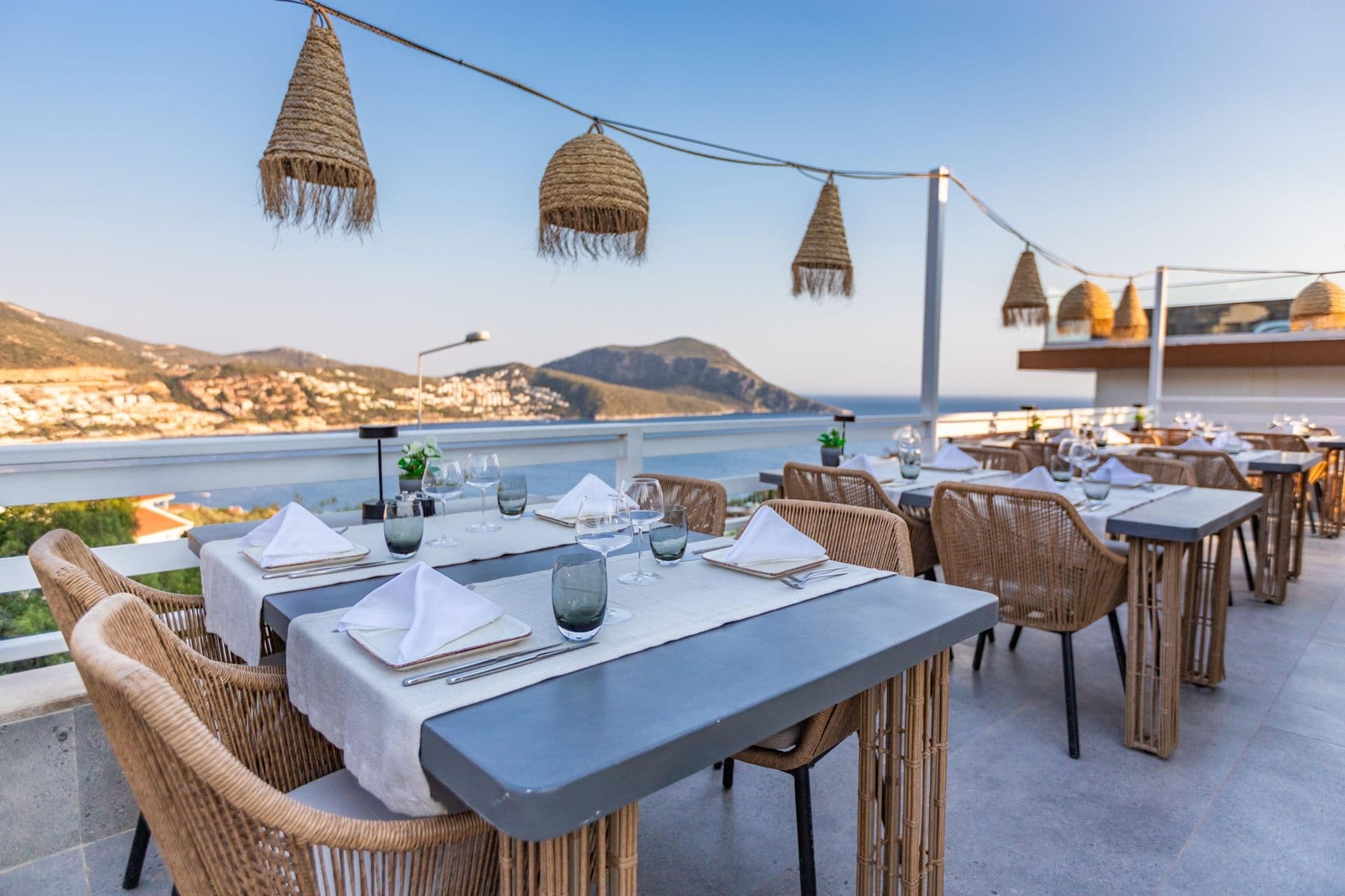 A rooftop terrace is set with tables for dining, string lights hung with woven shades, and hills and the sea beyond.