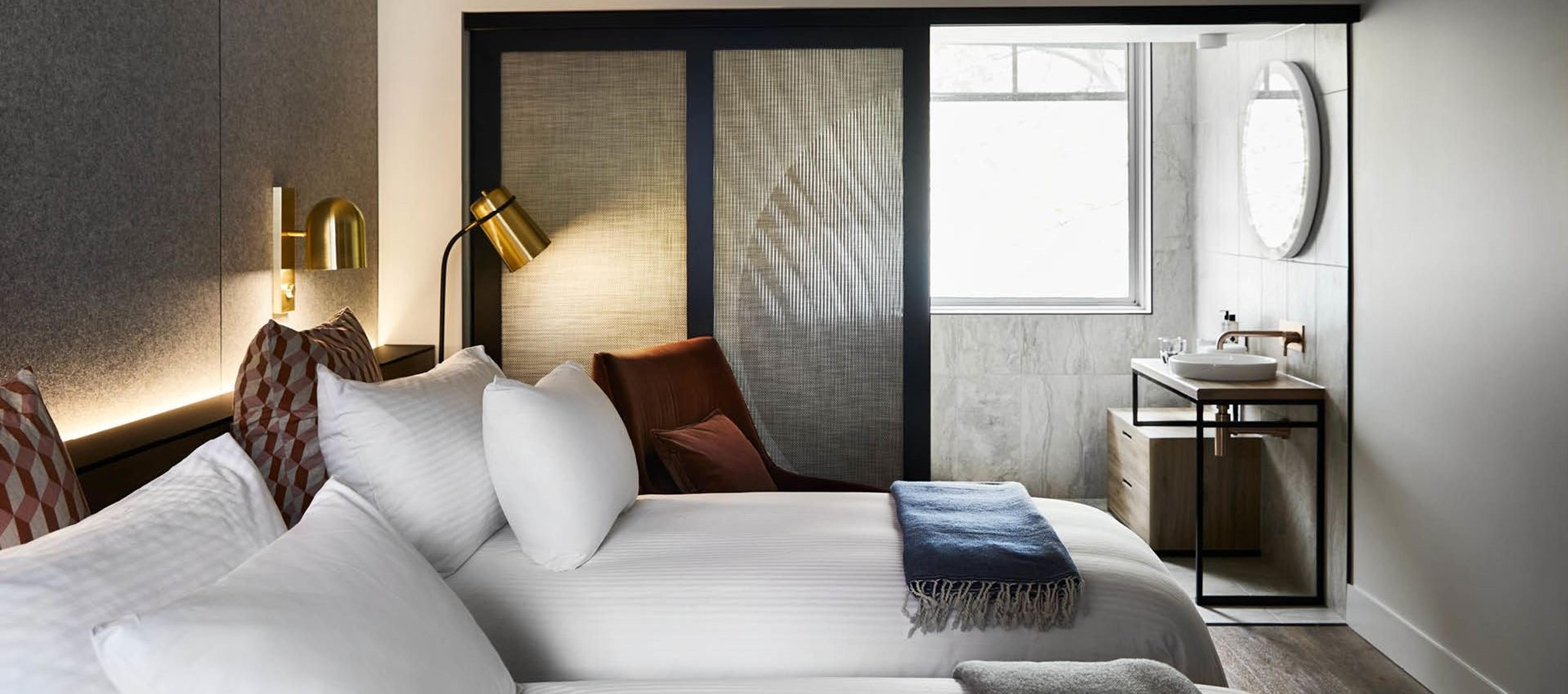 a twin hotel room with two beds, a burgundy armchair by the window, and a bathroom sink visible through an open screen