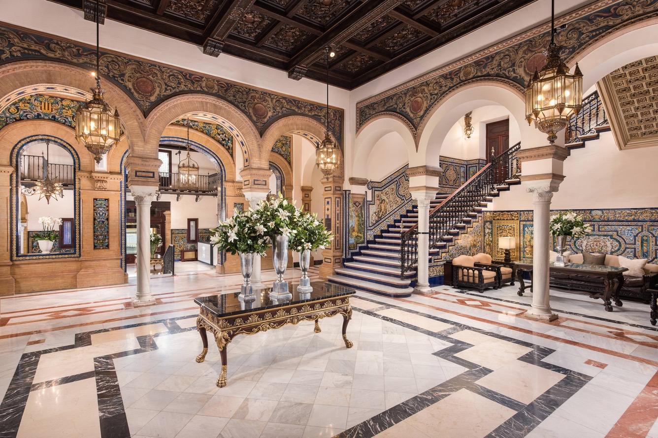 A grand tiled atrium with a carved wood ceiling, marble columns, a staircase, sofas and a large flower arrangement on a central table.