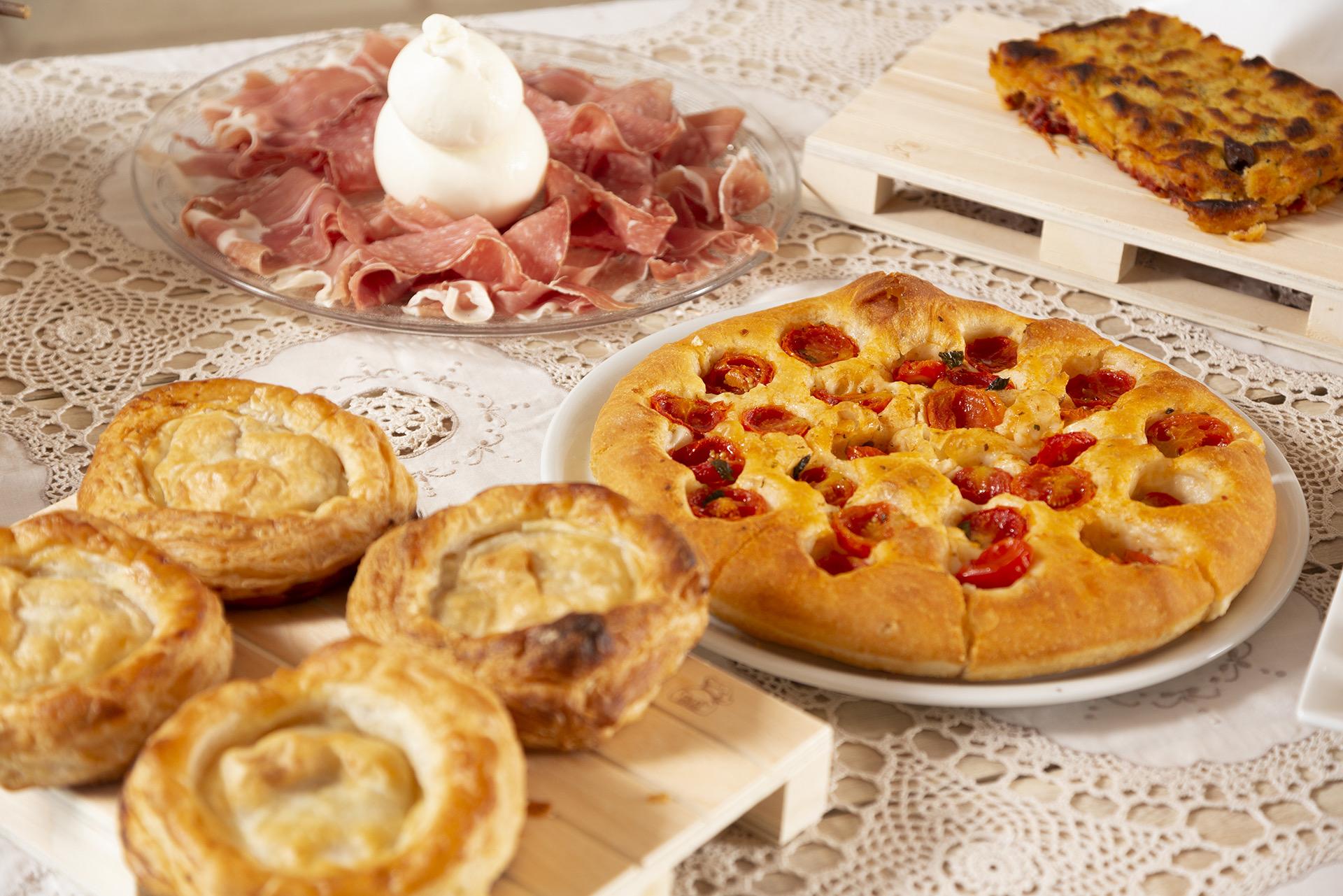 Sliced prosciutto and burrata, a tomato focaccia and small pastries laid out on a lace tablecloth.