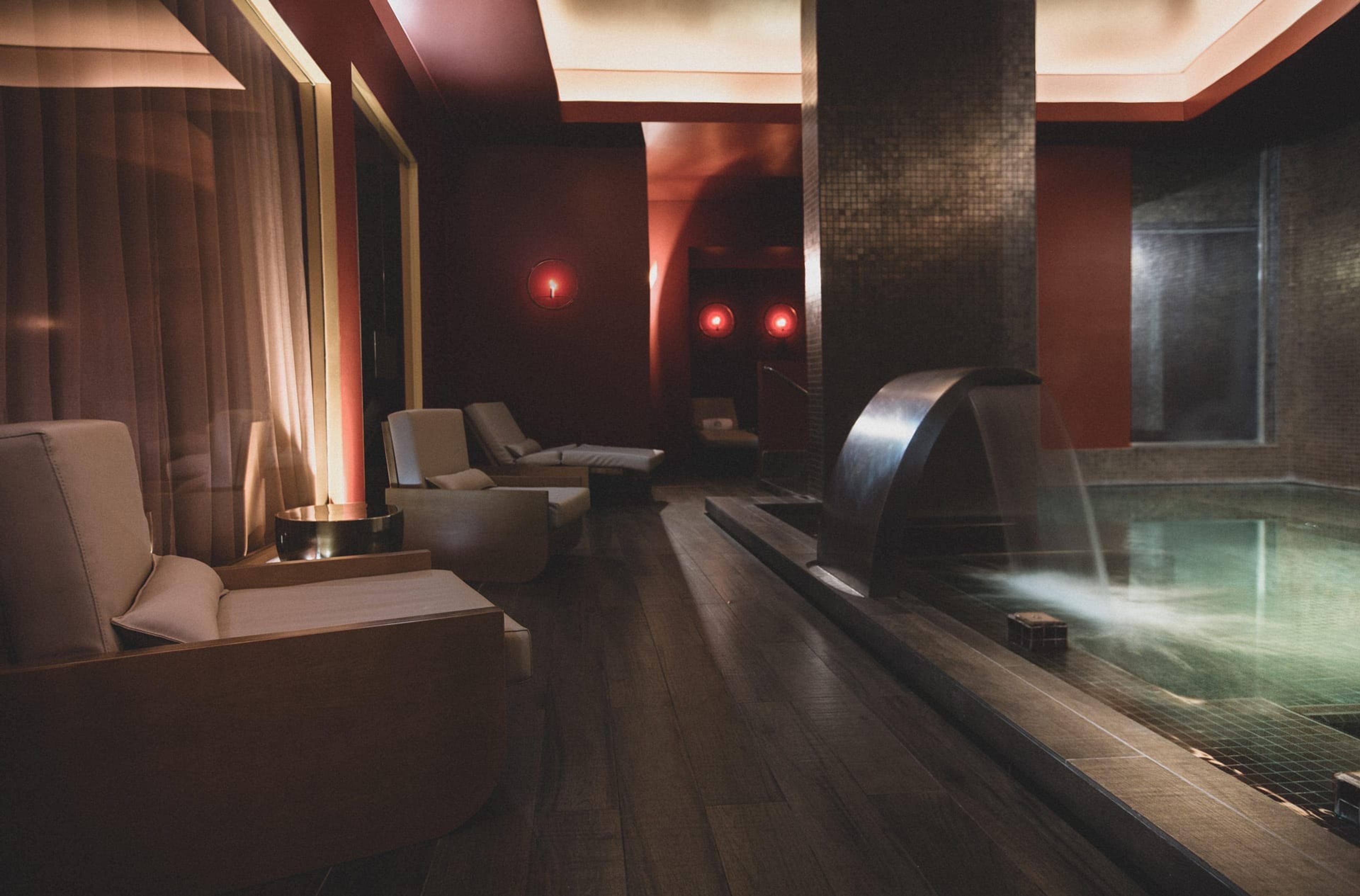An indoor pool with a dark tiled wall and a metal waterfall spilling into the water, with armchairs on a dark wooden floor under red light.