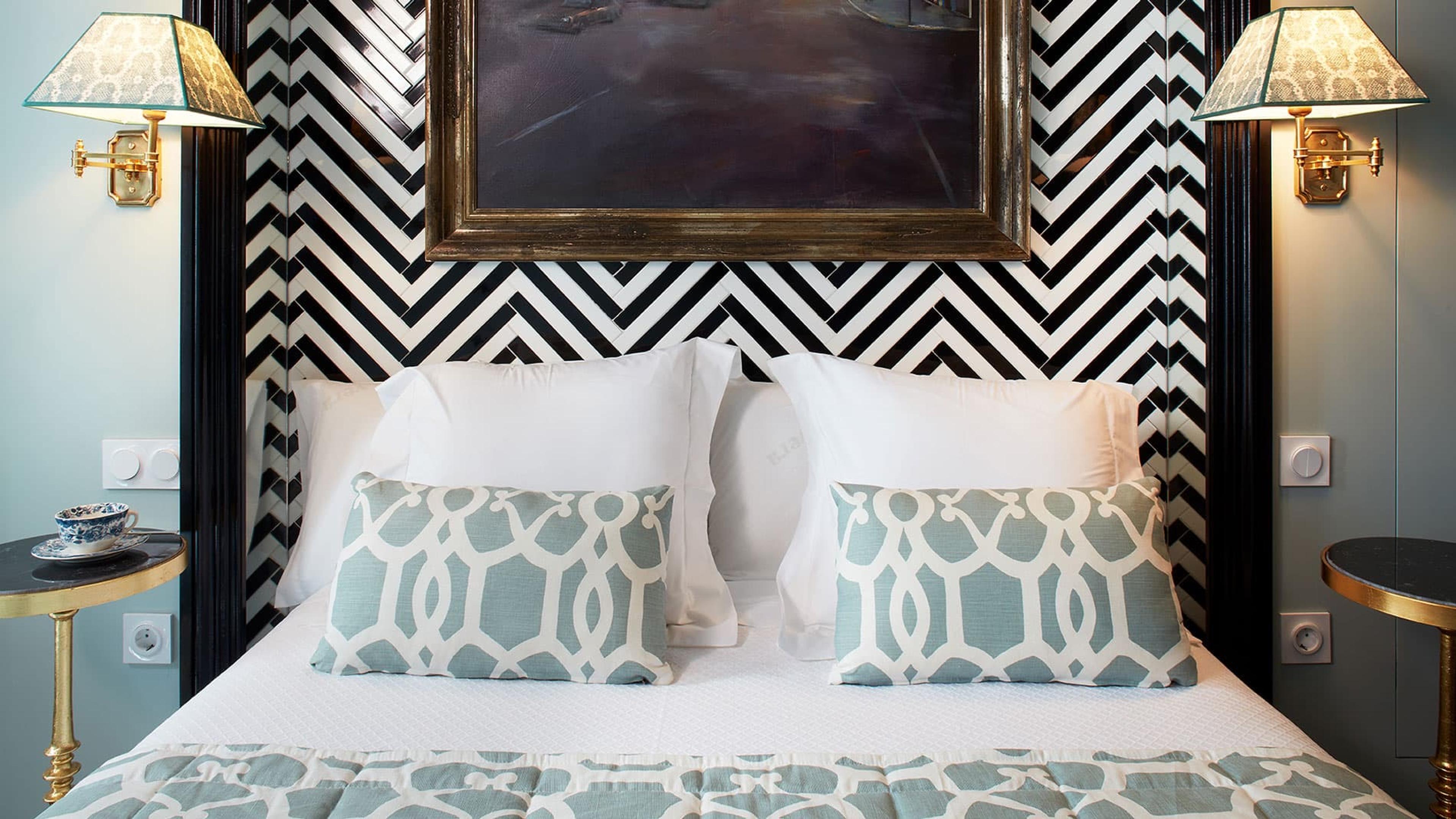 A made bed with a black and white chevron patterned headboard, teal patterned cushions, and a framed dark painting above.