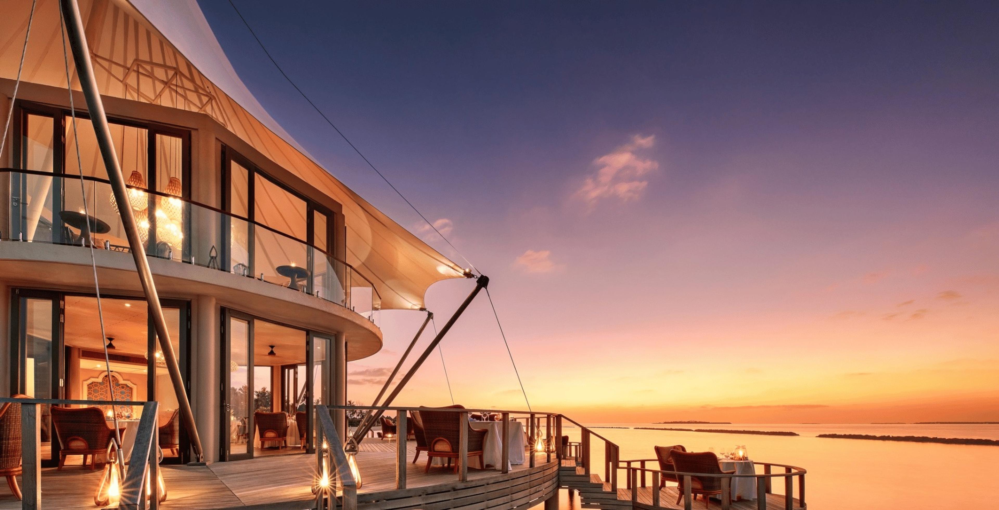A sail-shaped overwater restaurant lit from within at sunset, with tables set on its deck overlooking the ocean.