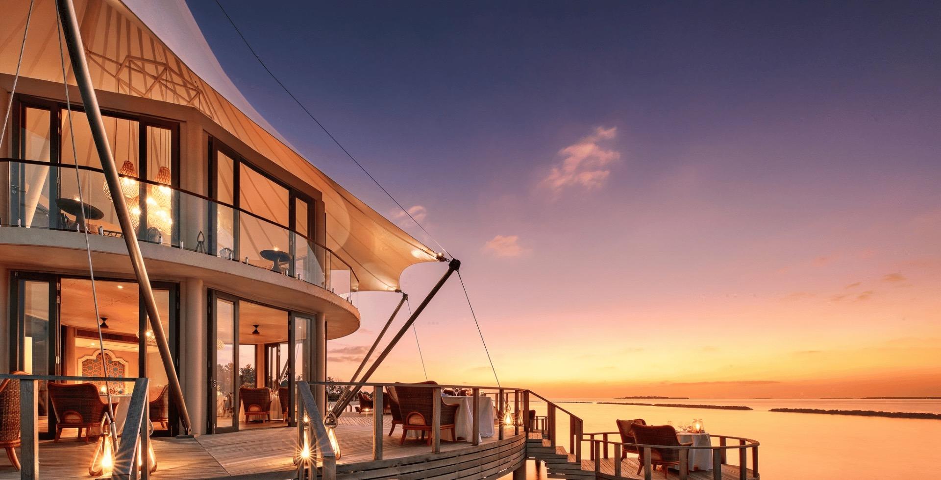 A sail-shaped overwater restaurant lit from within at sunset, with tables set on its deck overlooking the ocean.