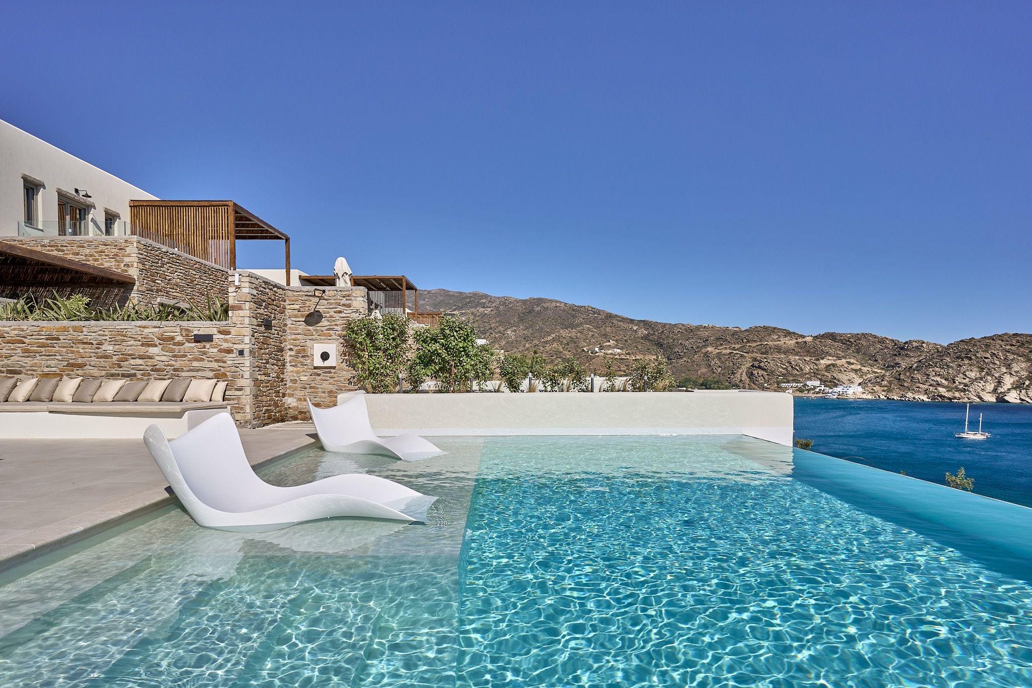 A pool with two white loungers floating in its shallow end, a stone-walled terrace behind, and hills and a sailing boat across the bay.