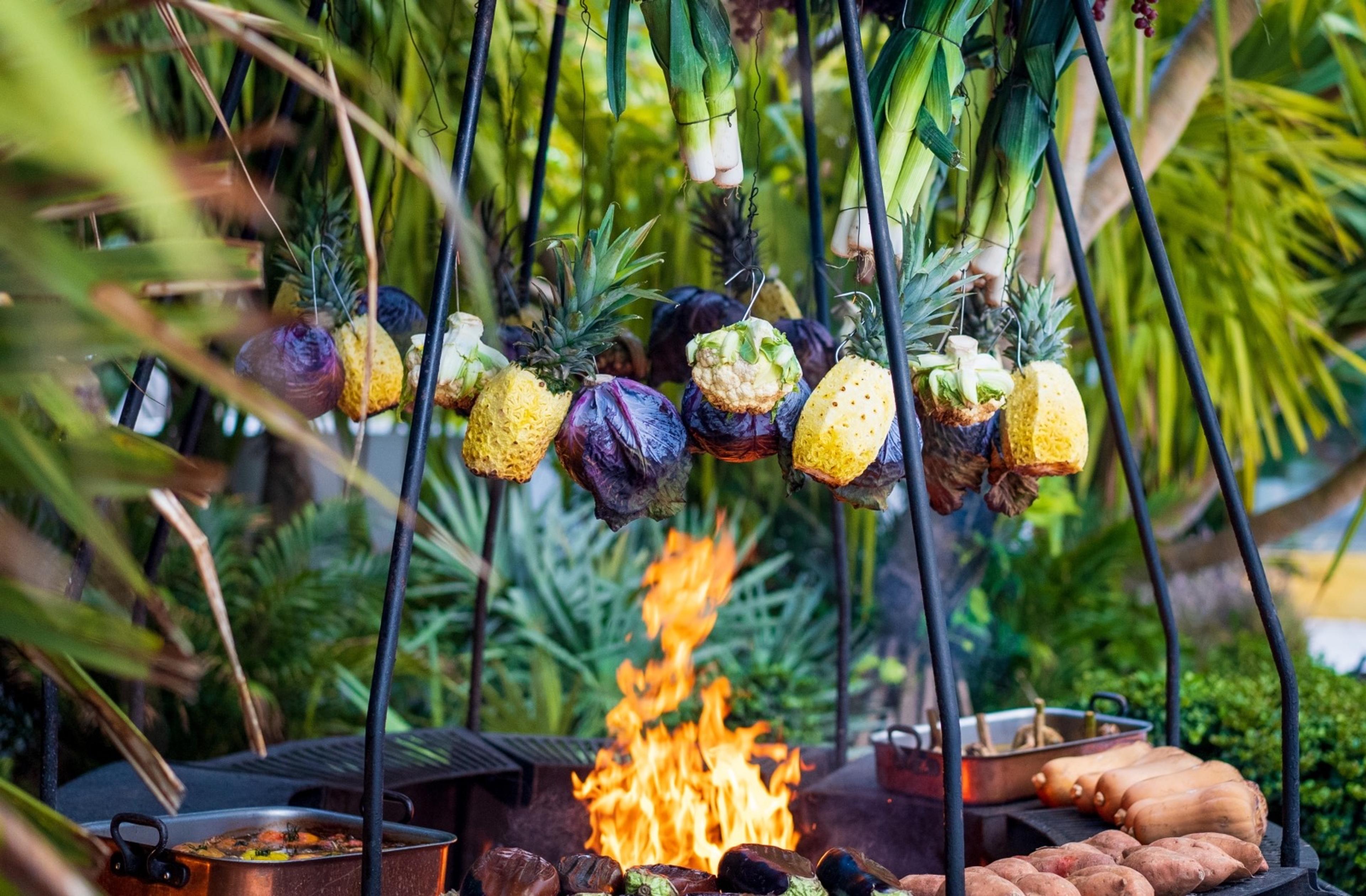 Pineapples, cabbages and corn hanging from iron spits over an open fire, with copper pans and trays of sweet potatoes beside it.