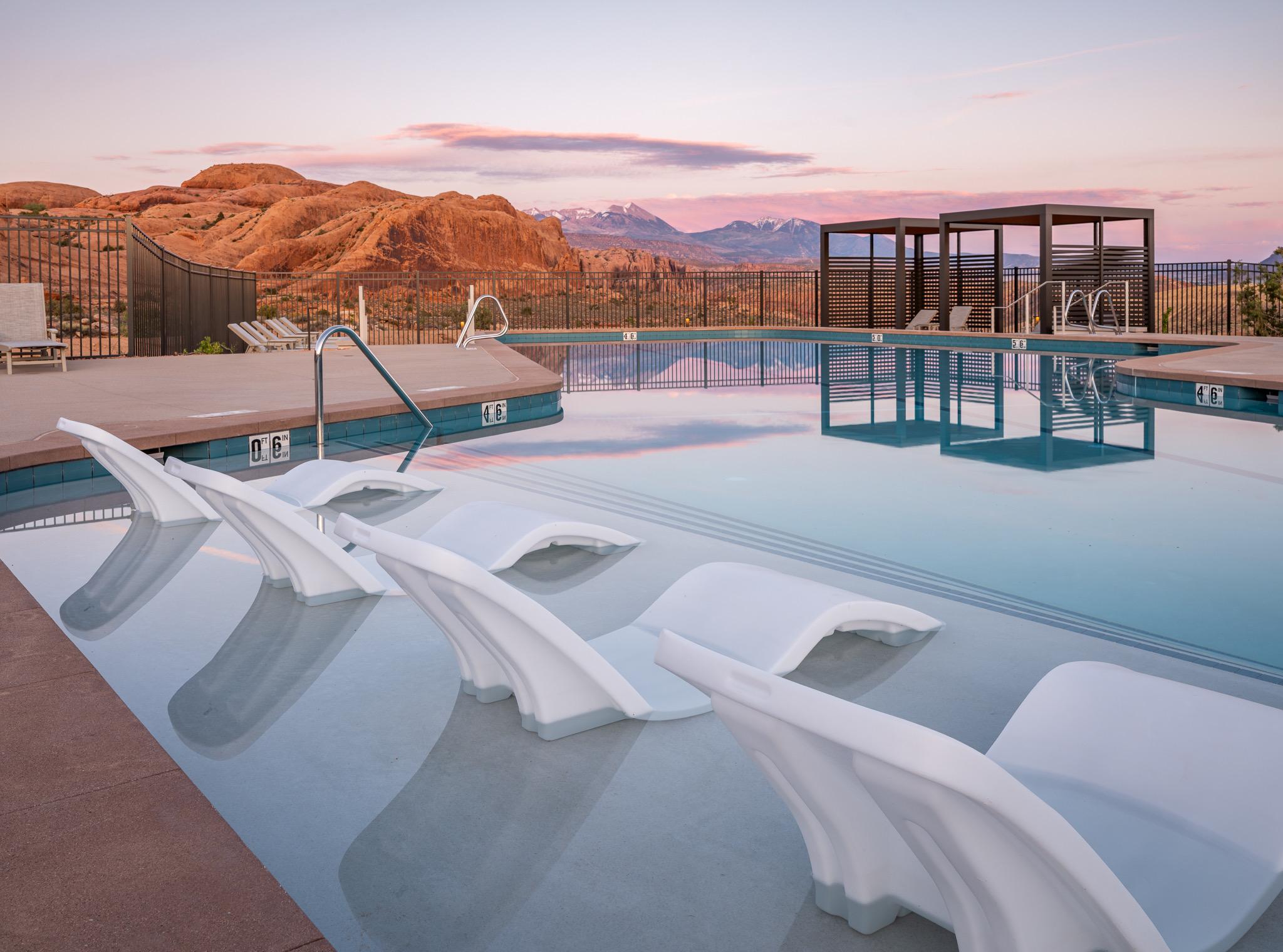 White lounge chairs sit submerged at the shallow end of a pool at dusk, with red rock cliffs and mountains beyond a metal fence.