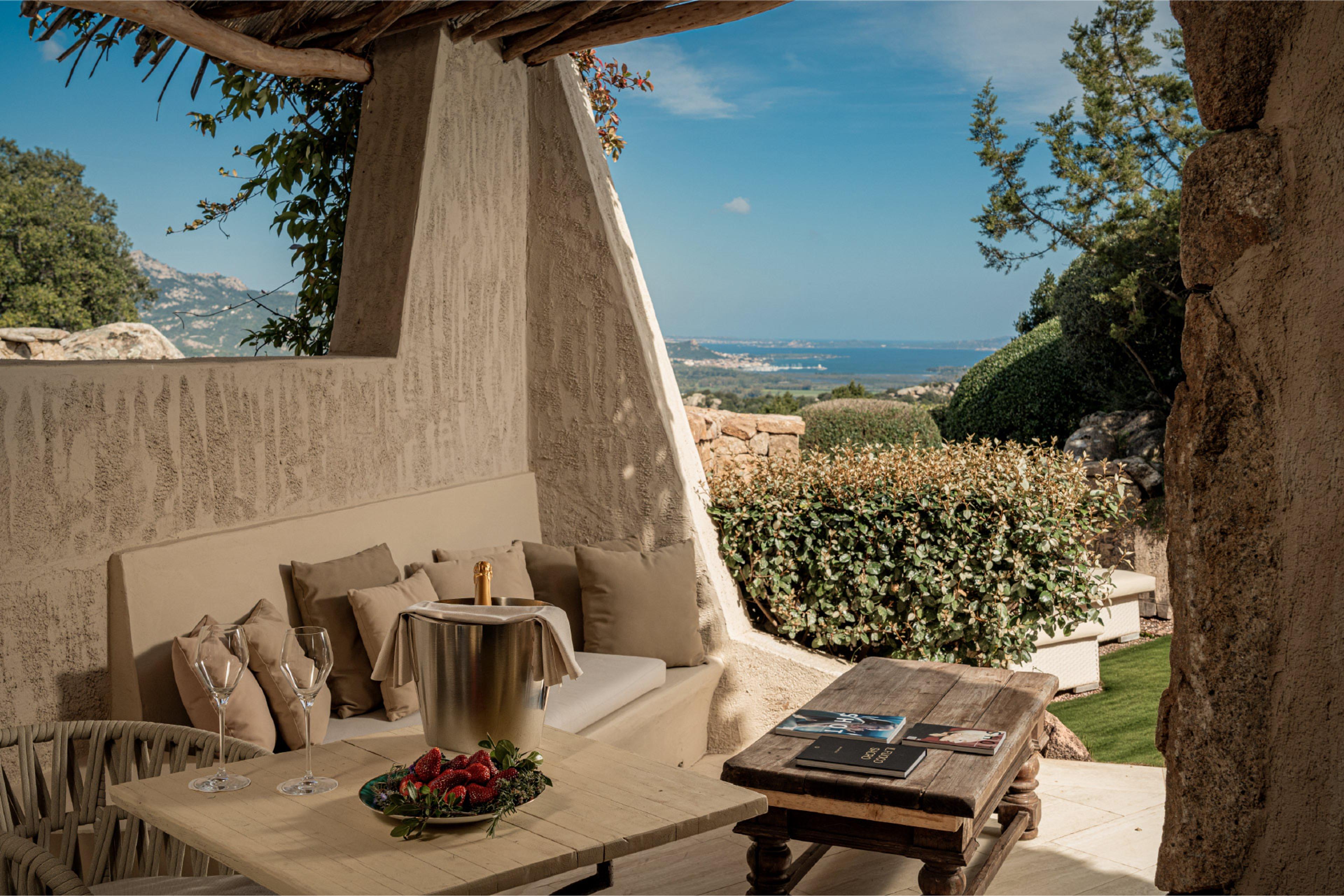 A terrace table laid with strawberries, wine glasses and a champagne bucket, with a cushioned bench and a sea view through pine trees.
