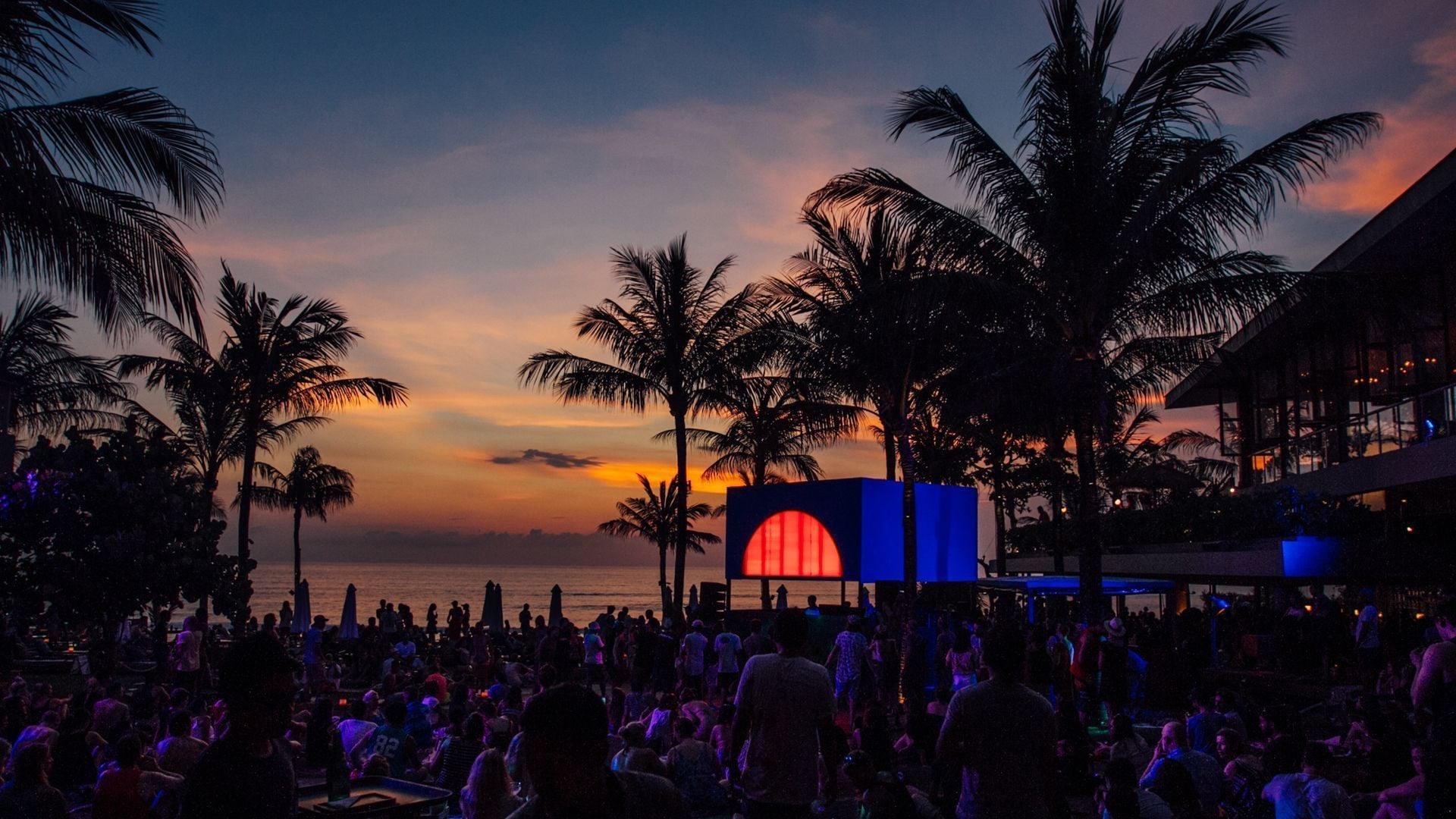 Silhouetted palm trees and a crowd gather on the beach at sunset beside a glowing stage box, the ocean turning orange behind them.