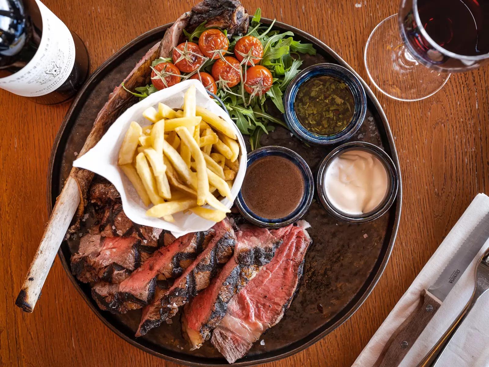 a sliced tomahawk steak on a plate with a bucket of chips, roasted tomatoes, three sauces, a glass of red wine and a bottle alongside