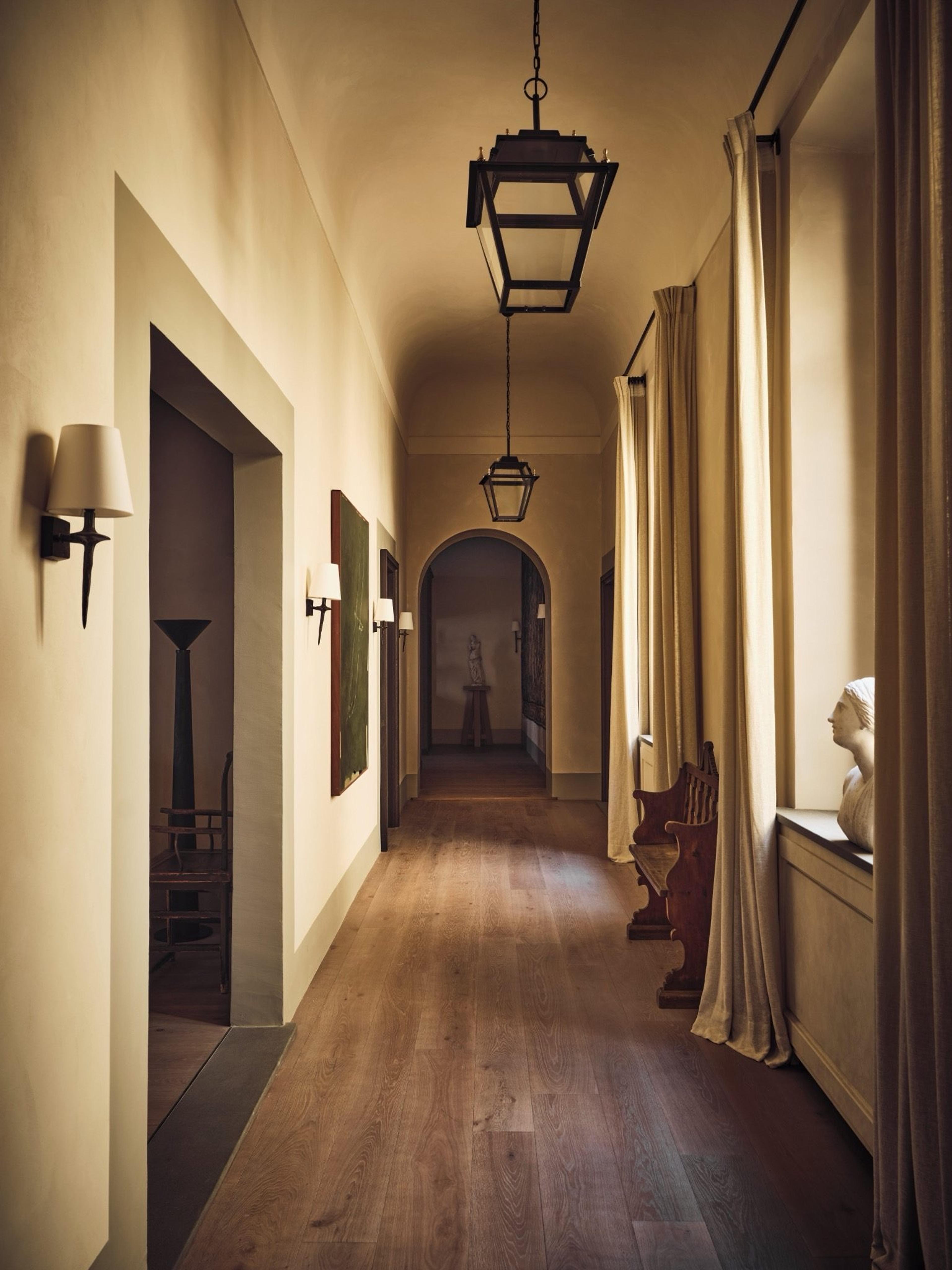 A long vaulted corridor with hanging lantern light fixtures, a wooden bench against curtained windows, and a stone bust on a console table.