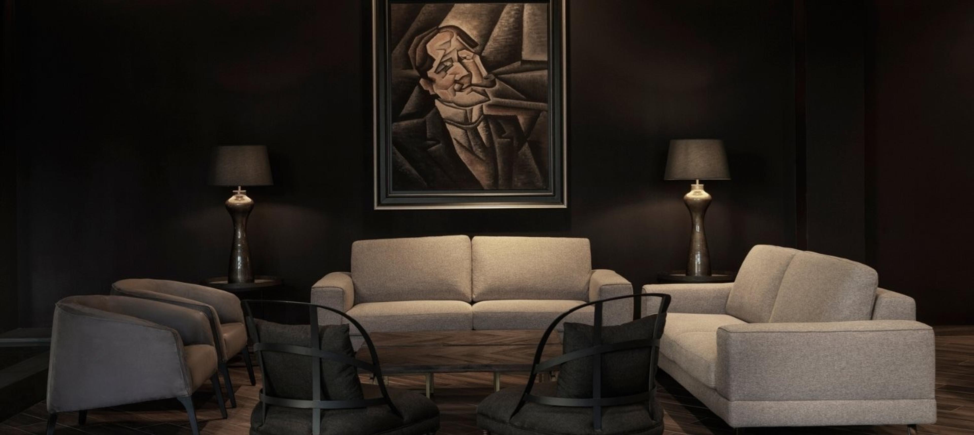 A dark lounge with beige sofas and chairs around a low table, a framed portrait and lamps on the wall behind.