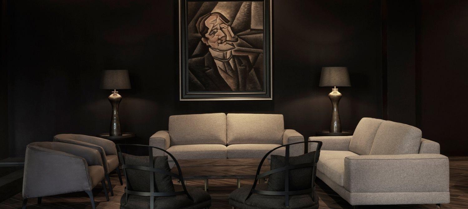 A dark lounge with beige sofas and chairs around a low table, a framed portrait and lamps on the wall behind.