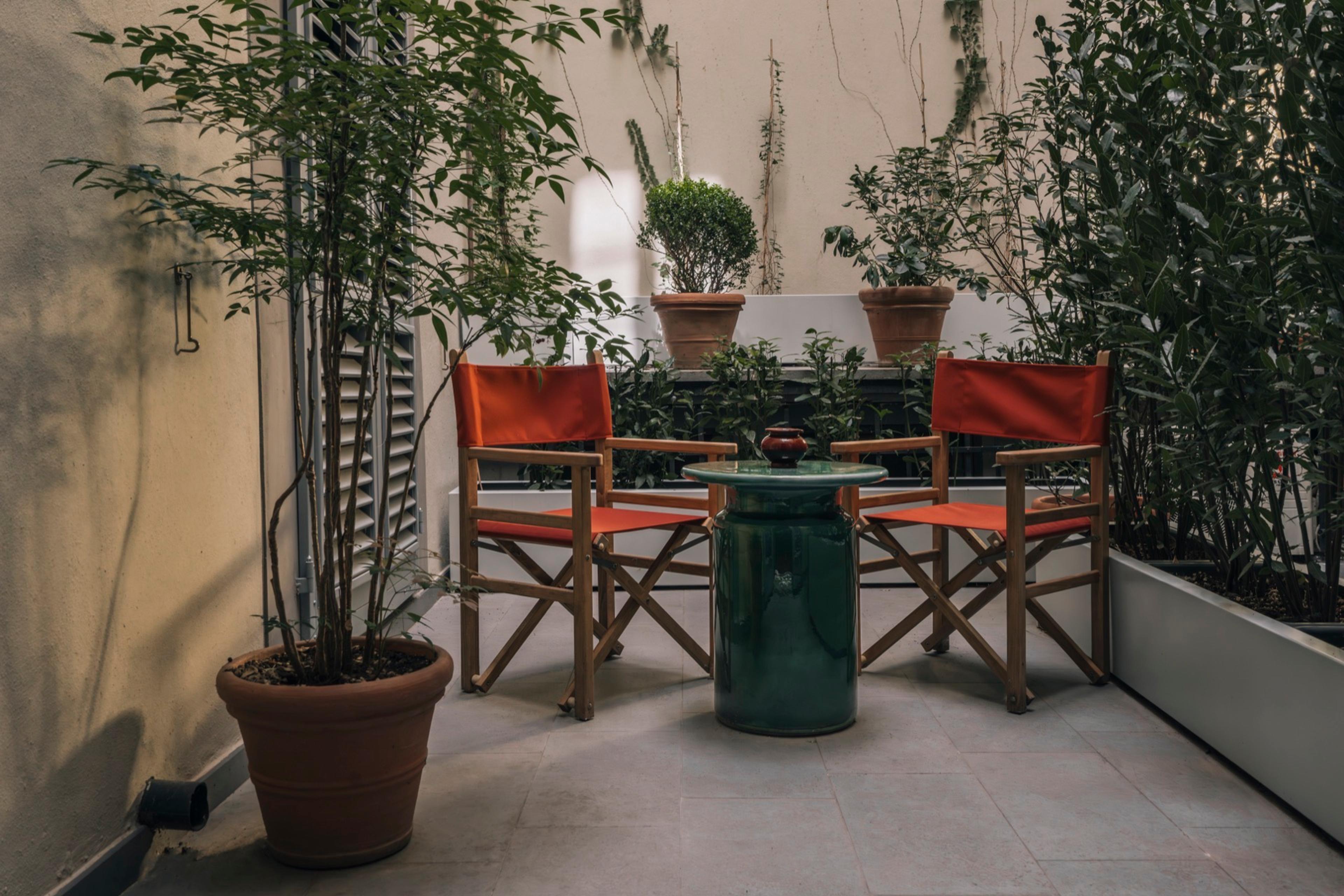 Two red director's chairs and a green ceramic side table sit on a tiled terrace crowded with potted plants against the building's wall.