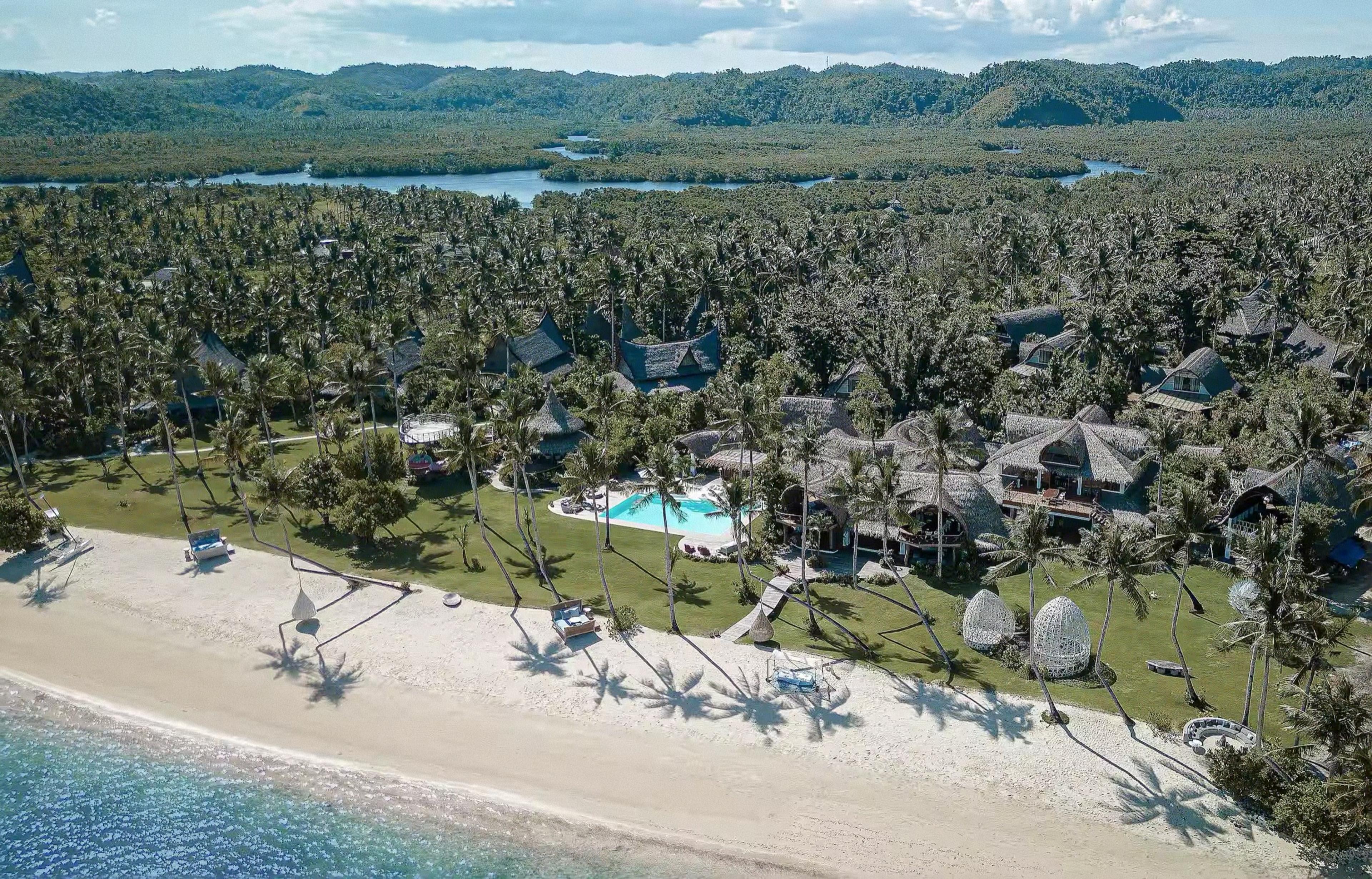 An aerial daylight view of a beachfront resort of thatched roof buildings and a pool among palm trees, with a sandy beach, sea and forested hills beyond.