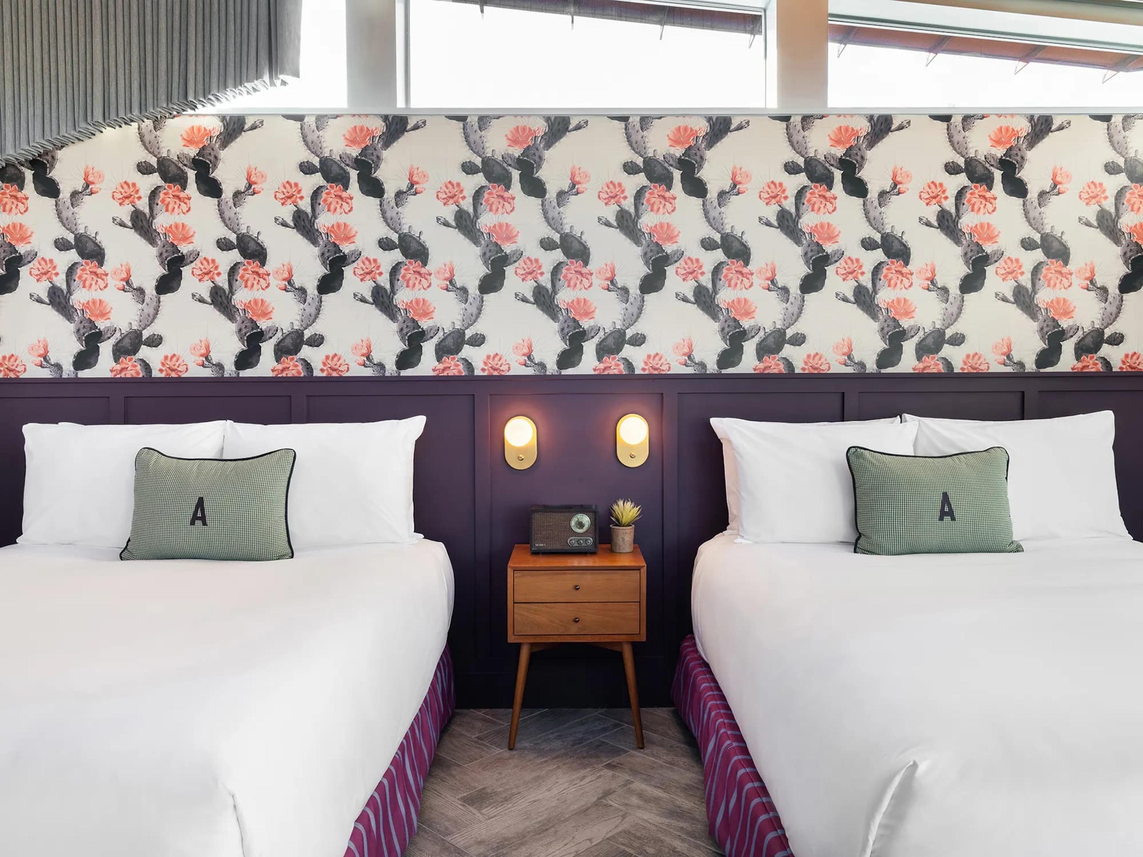 Two double beds with white bedding and green cushions marked with the letter A, in front of a dark plum panelled wall, with a cactus-print wallpaper band above and a wooden bedside table between them.