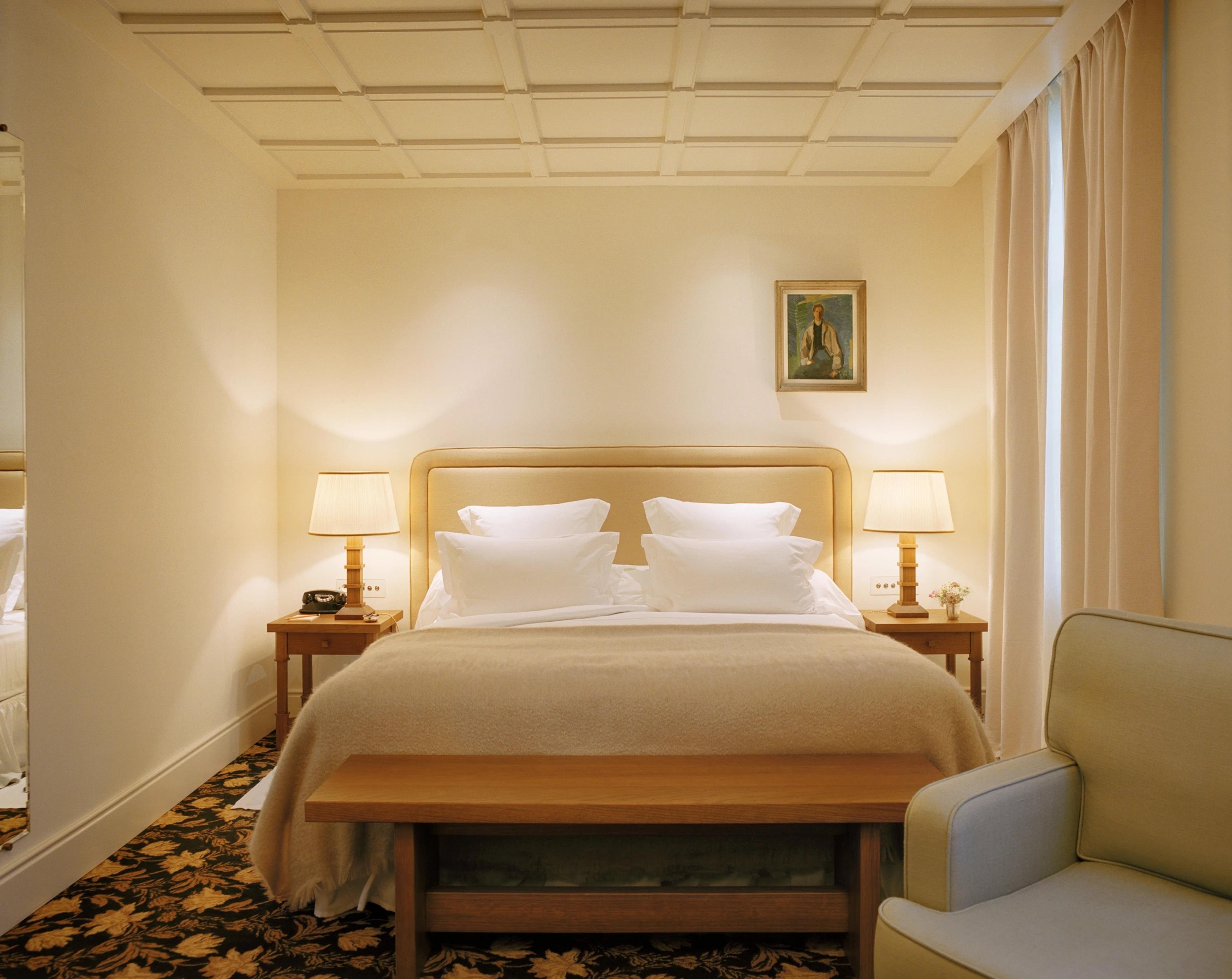 A bedroom in cream with a padded headboard, a bench at the foot of the bed and lamps either side.