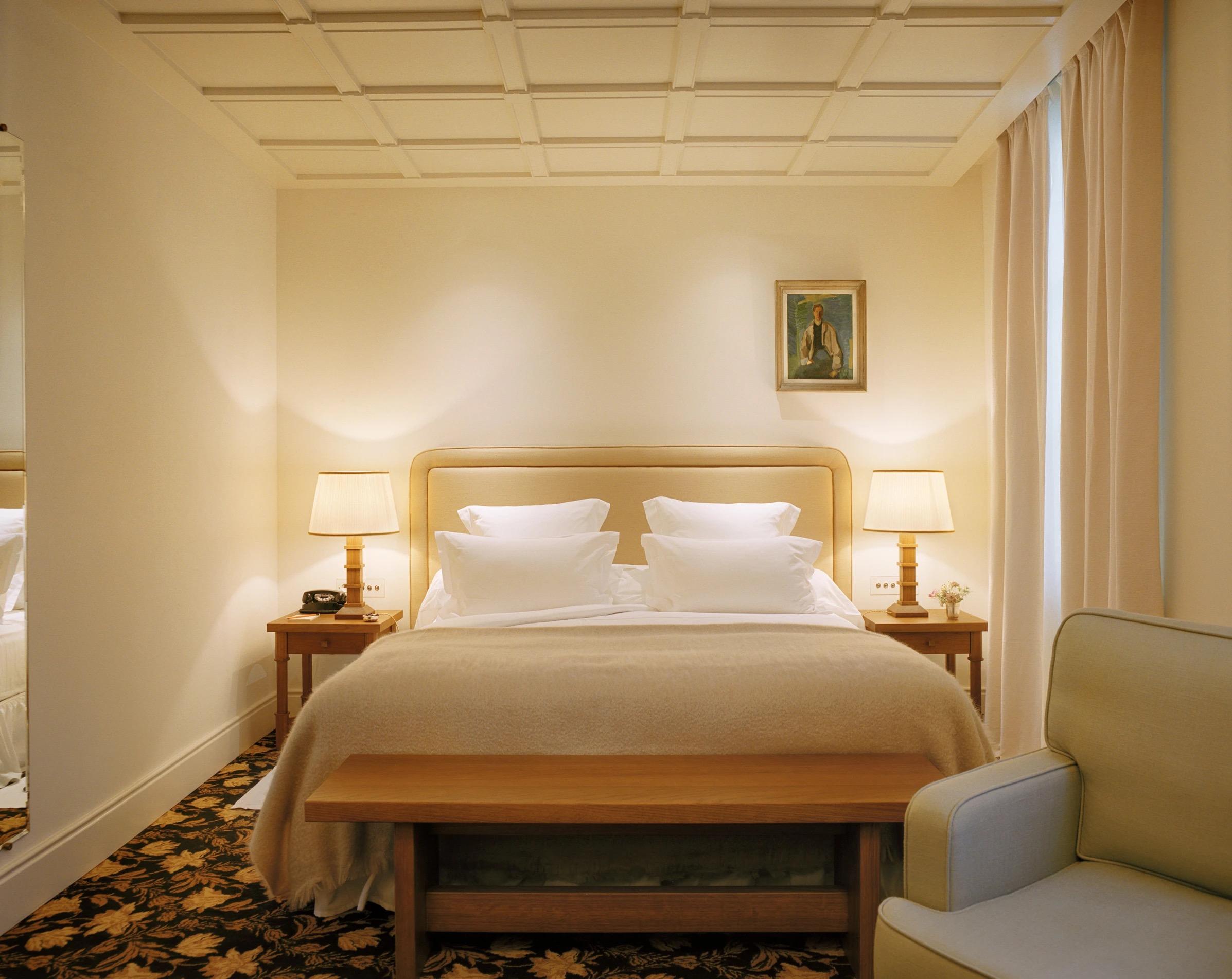 A bedroom in cream with a padded headboard, a bench at the foot of the bed and lamps either side.