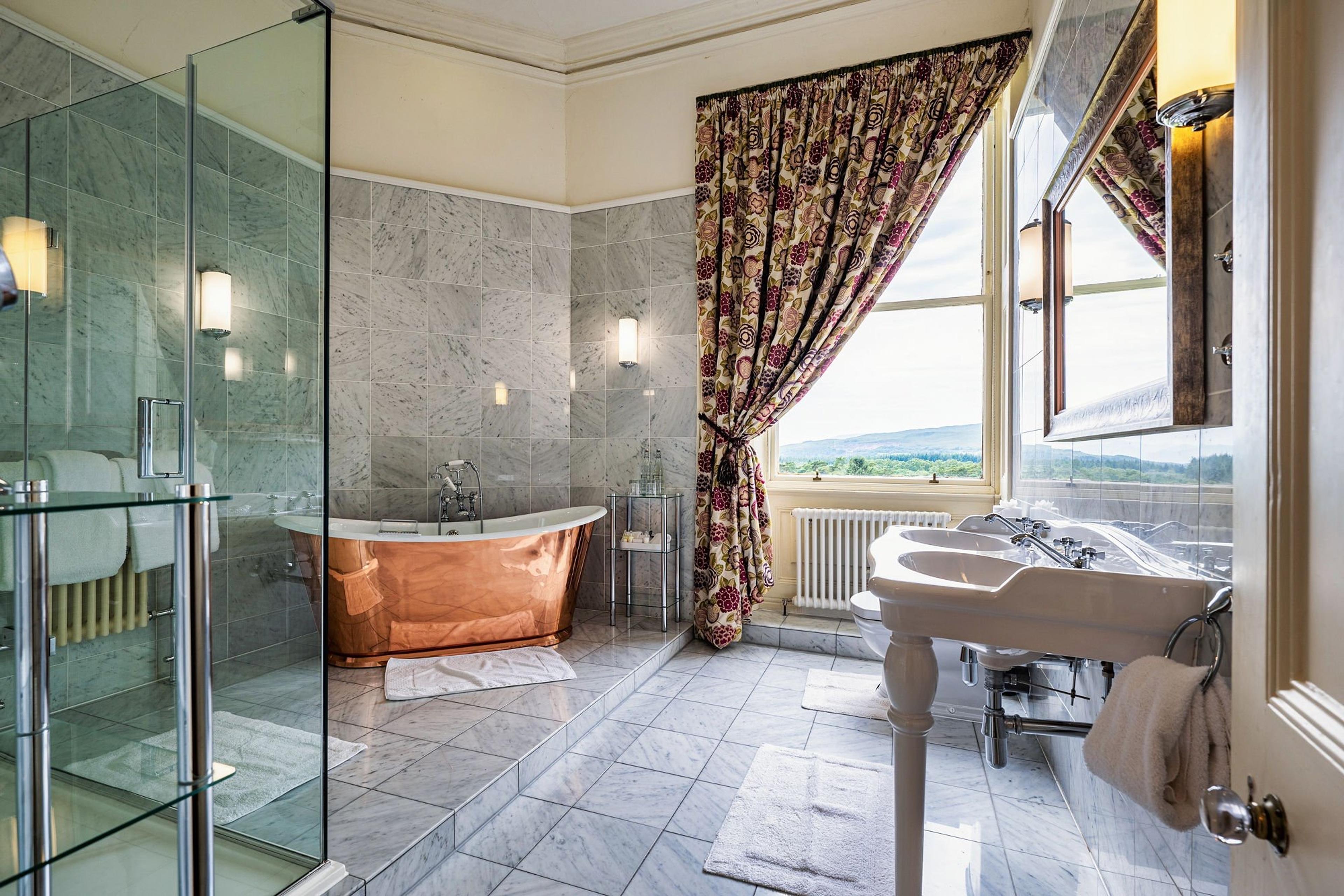 A marble bathroom with a free-standing copper bath, a glass shower, twin sinks and a curtained window looking over the hills.