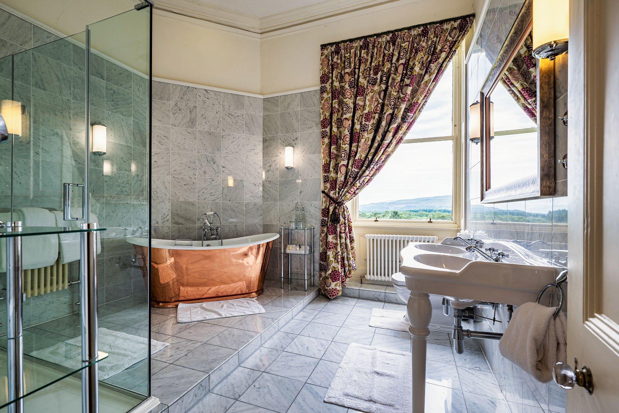 A marble bathroom with a free-standing copper bath, a glass shower, twin sinks and a curtained window looking over the hills.