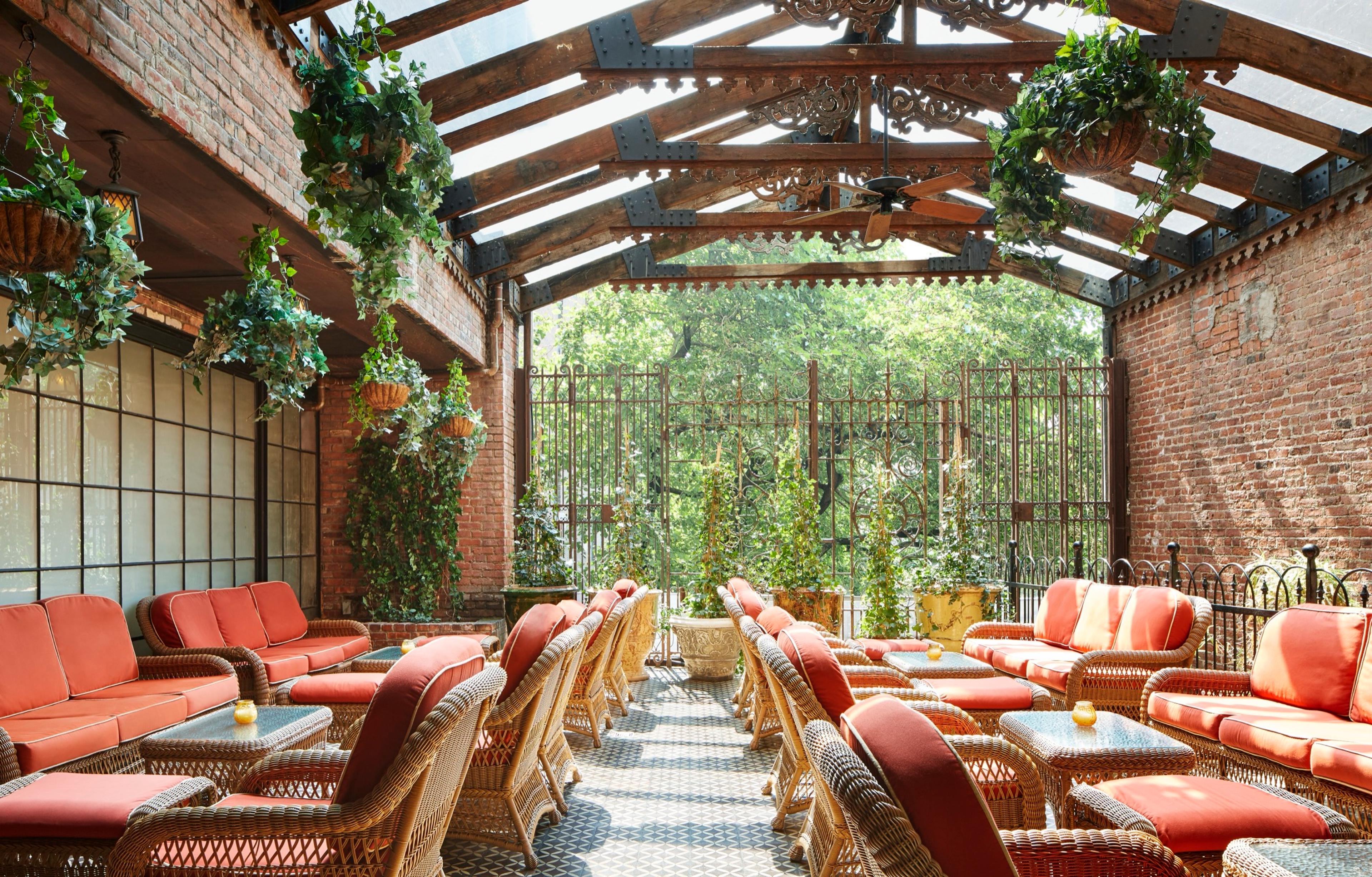 A glass-roofed garden patio with rows of wicker chairs and sofas in coral cushions, hanging plants, and trees visible through an iron gate.
