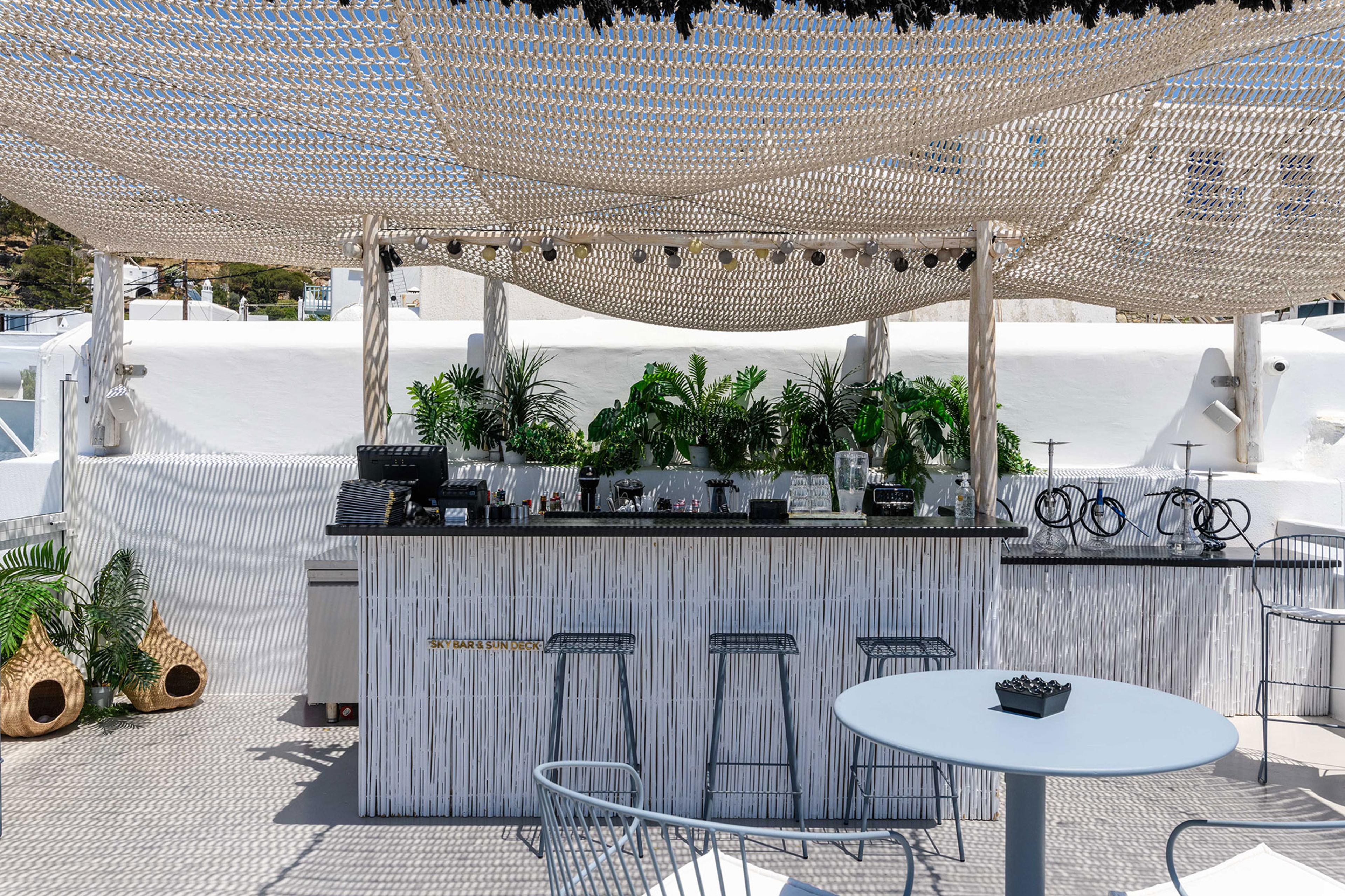 An outdoor bar with potted plants under a woven canopy, with stools and a round table on a terrace.