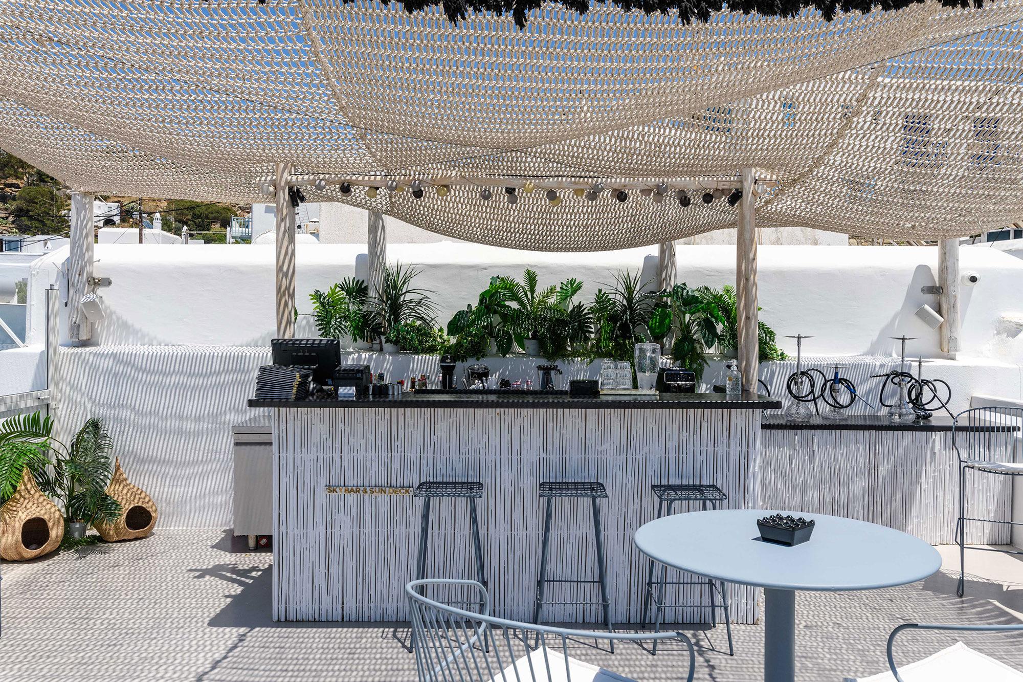 An outdoor bar with potted plants under a woven canopy, with stools and a round table on a terrace.