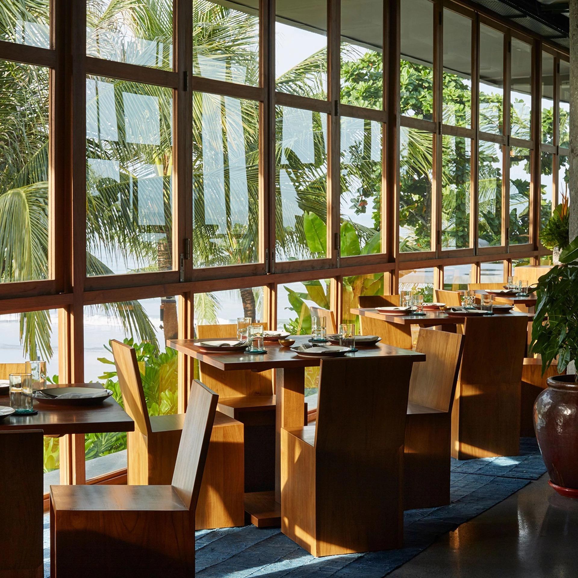 Empty wooden dining tables and chairs face floor-to-ceiling windows looking onto palm trees and the sea.