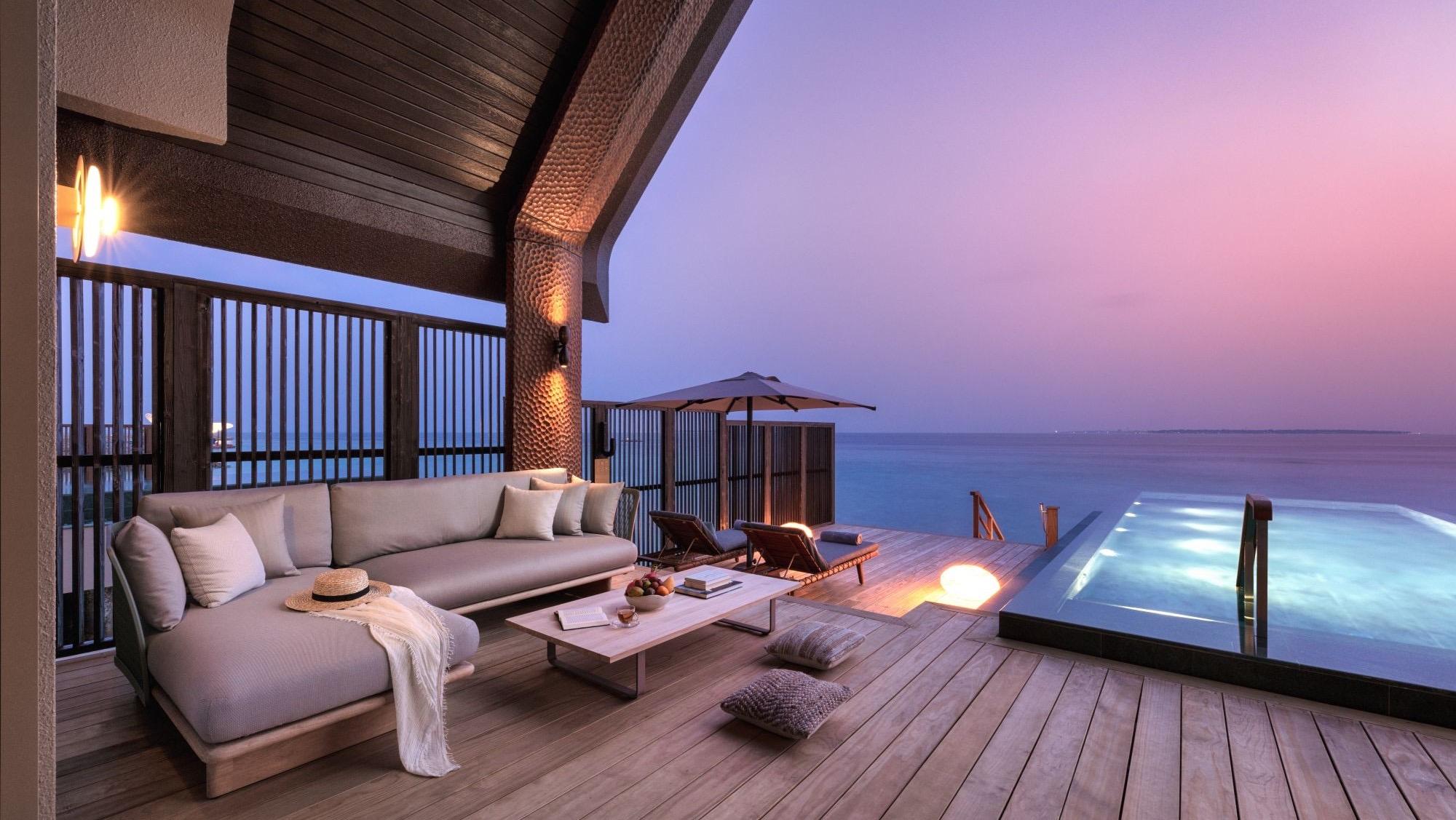 An over-water villa deck with a sofa, a low table and lanterns, its pool running out towards the sea at sunset.