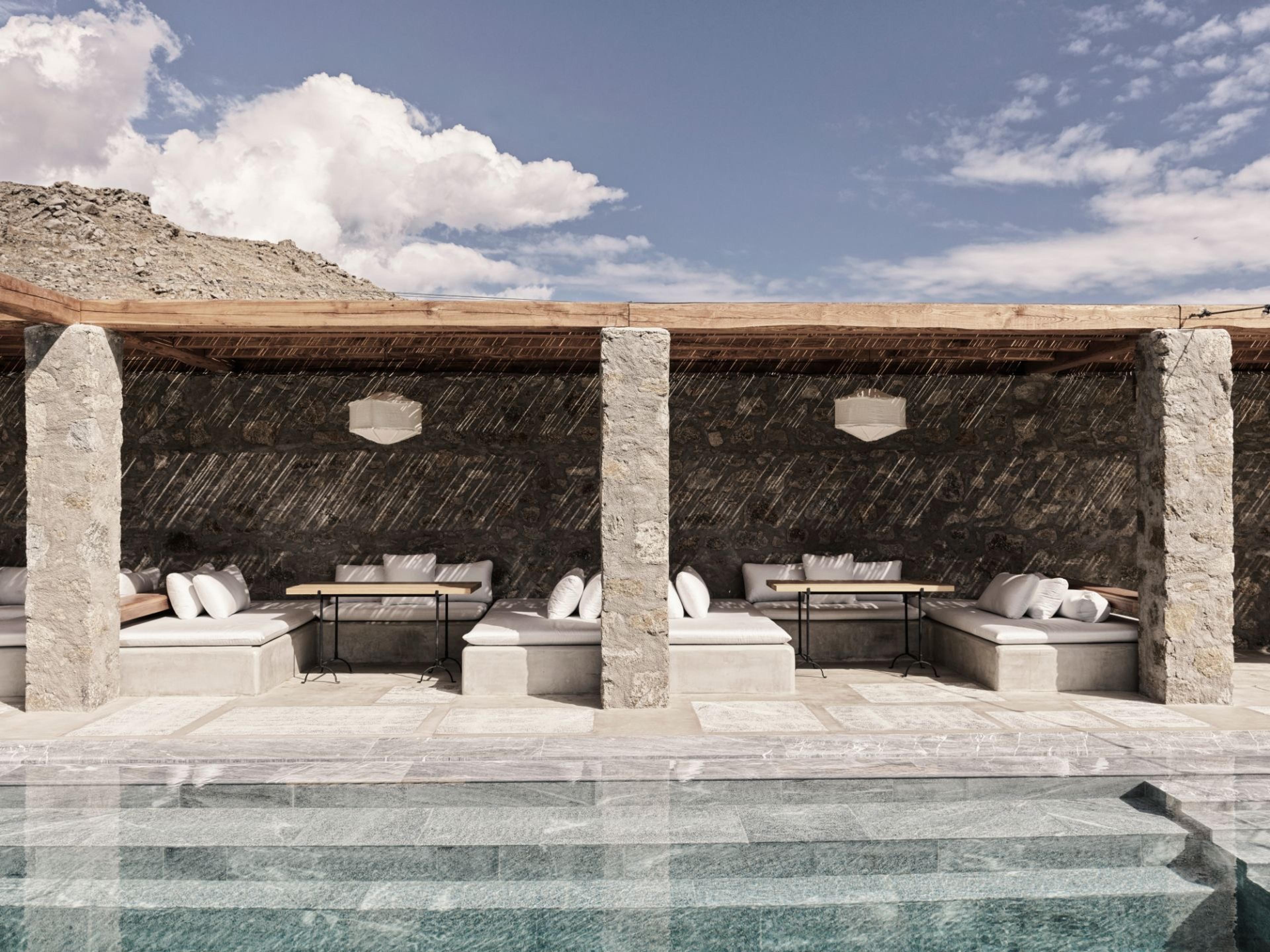 A row of stone-pillared daybeds sits under a wooden pergola beside a pool, with a rocky hillside and scattered clouds behind.