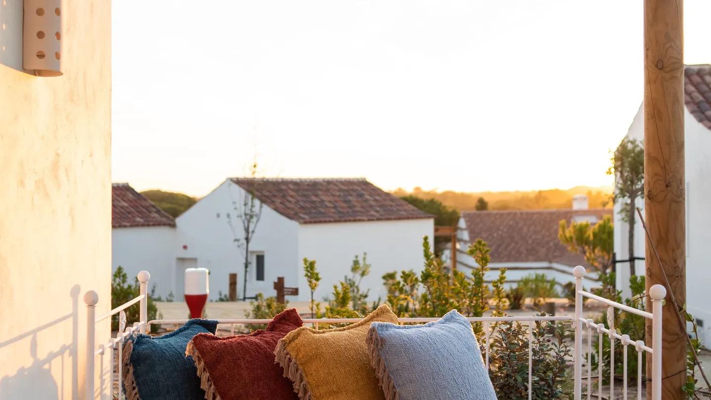 four tasseled cushions lined along a white iron railing on a terrace, with white village houses and a rooftop view behind at sunset