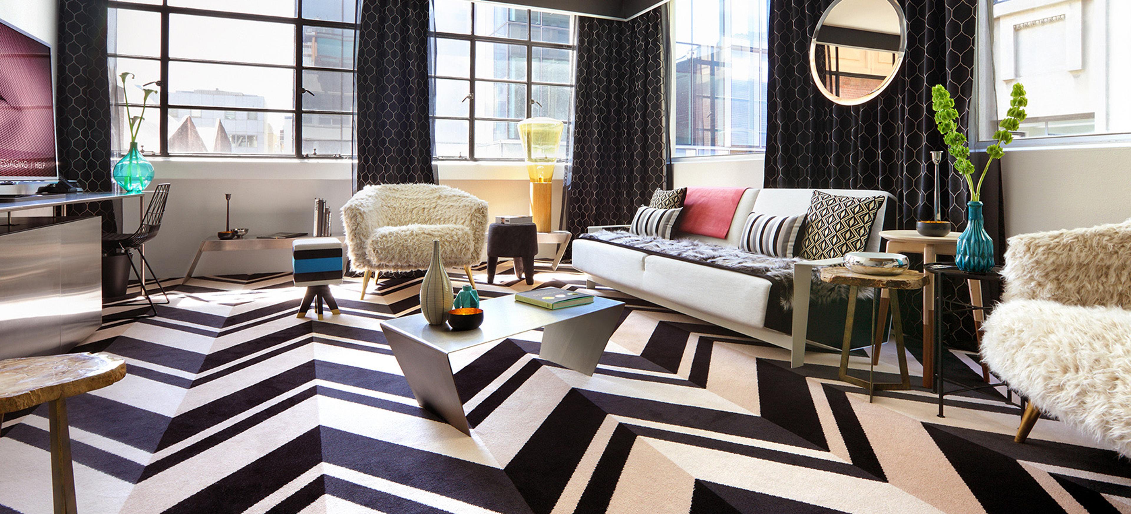 A bright living room with a white sofa and shaggy armchairs around a steel coffee table, tall windows looking onto the street, and a round mirror on a patterned wall.