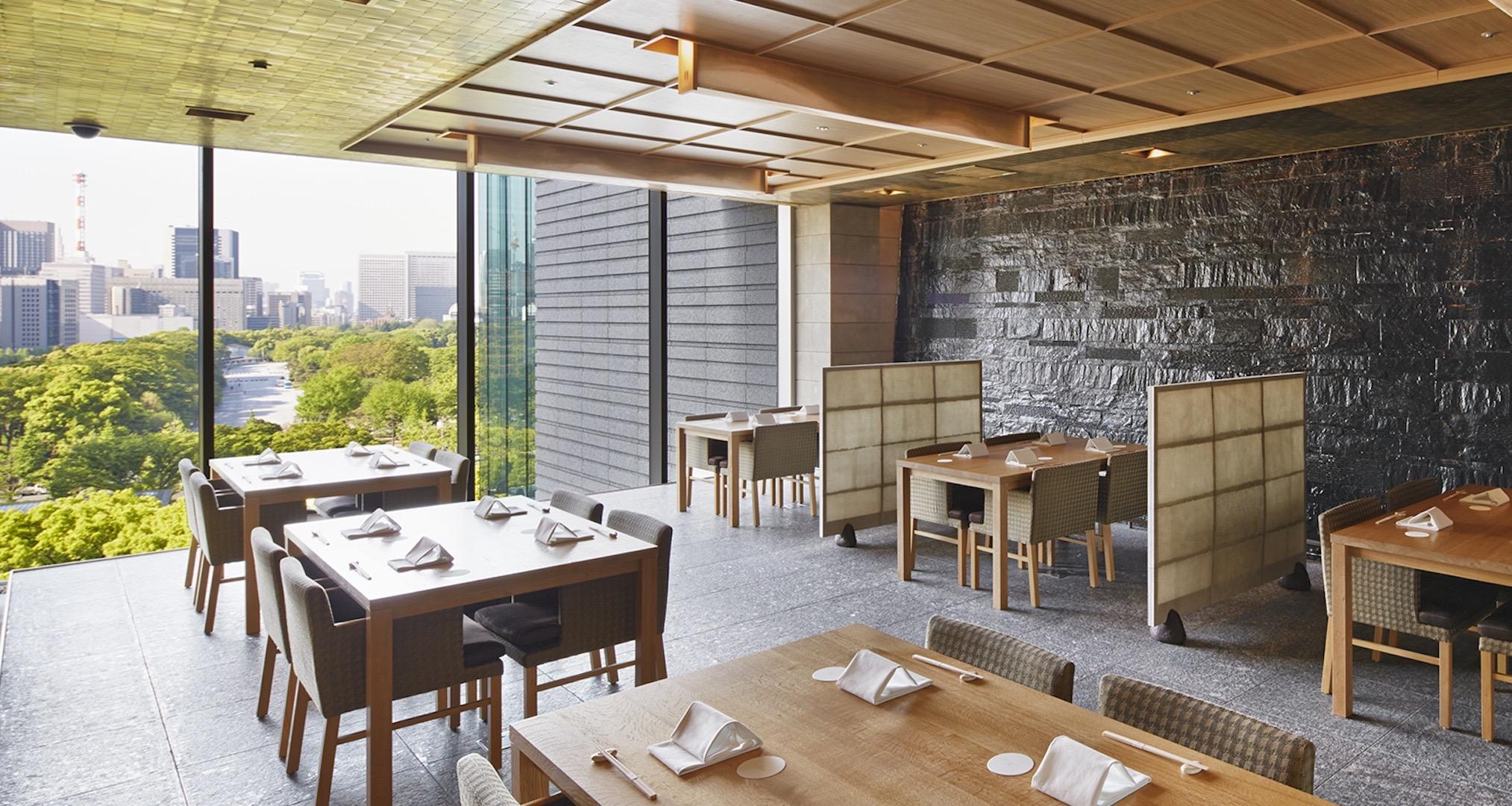 A restaurant dining room with tables set for a meal, woven chairs, a dark stone wall and glass looking out over treetops to the skyline.