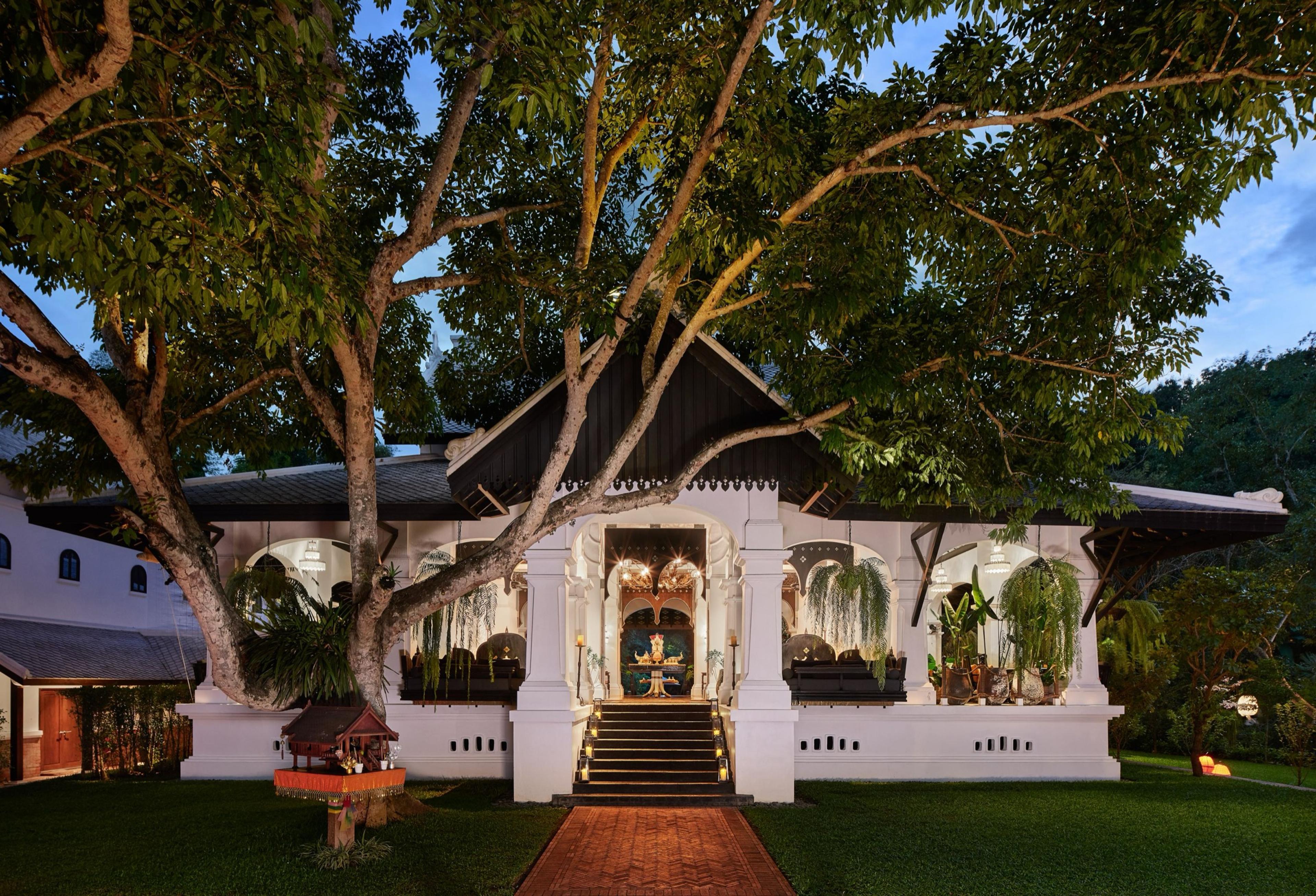 A white colonial-style building with a dark gabled roof, lit from within at dusk, with a large spreading tree in front and a brick path leading to the entrance steps.