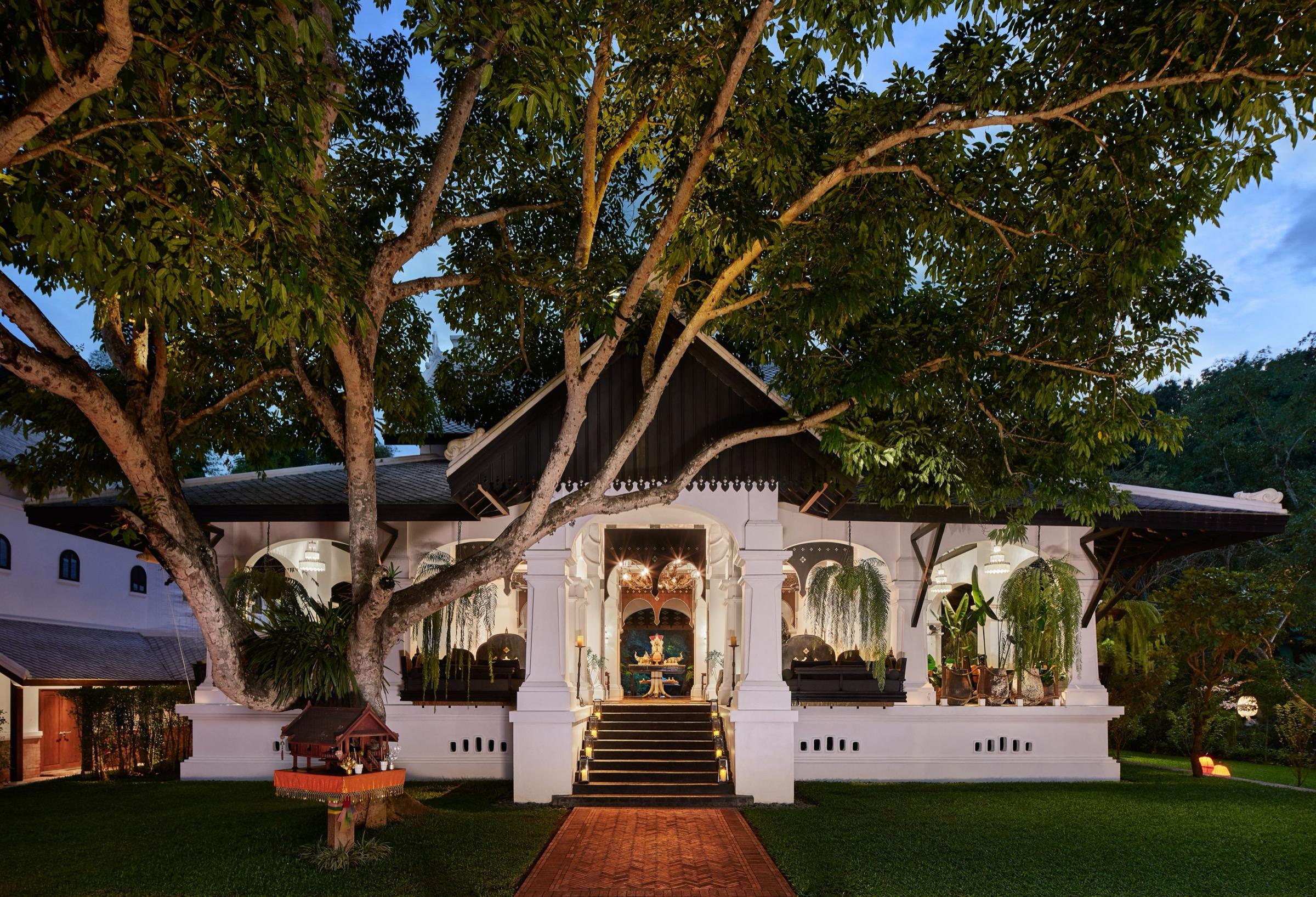 A white colonial-style building with a dark gabled roof, lit from within at dusk, with a large spreading tree in front and a brick path leading to the entrance steps.