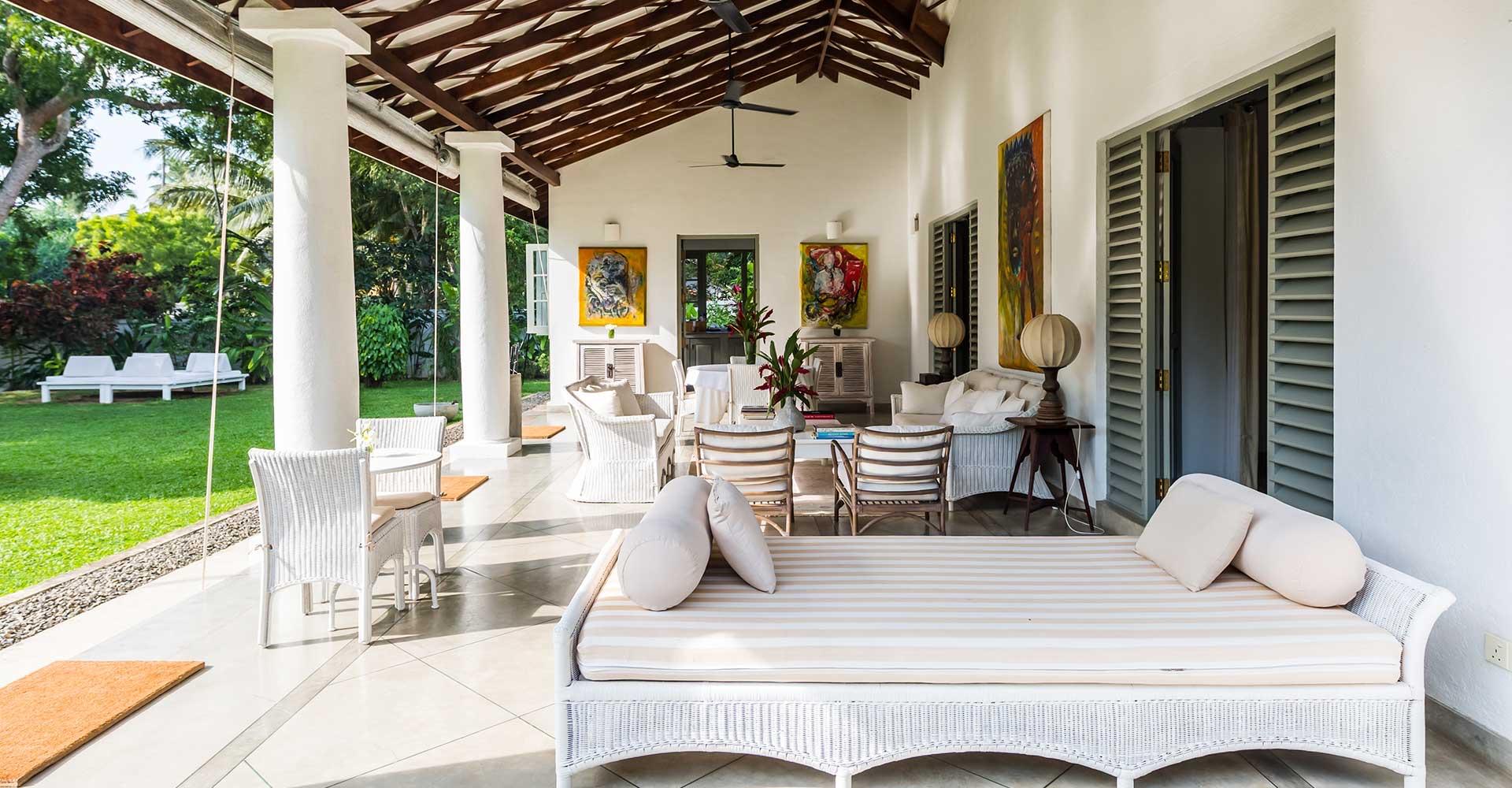 A covered veranda with a striped daybed and cushions, a timber-beamed roof and white columns, with wicker chairs and the lawn beyond.
