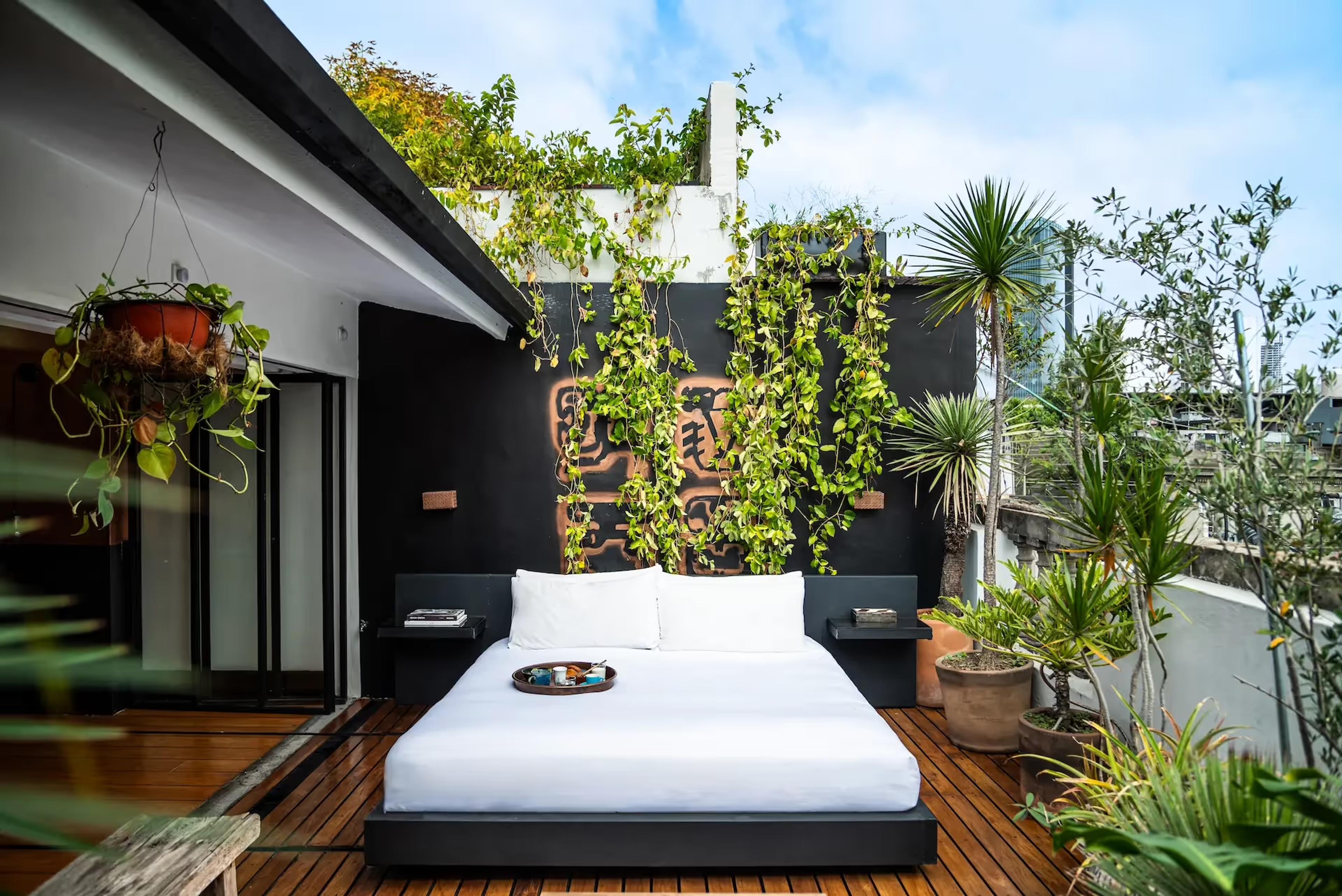 A black platform bed sits on a wooden rooftop deck against a black wall hung with trailing vines and a copper mural, city buildings visible beyond.