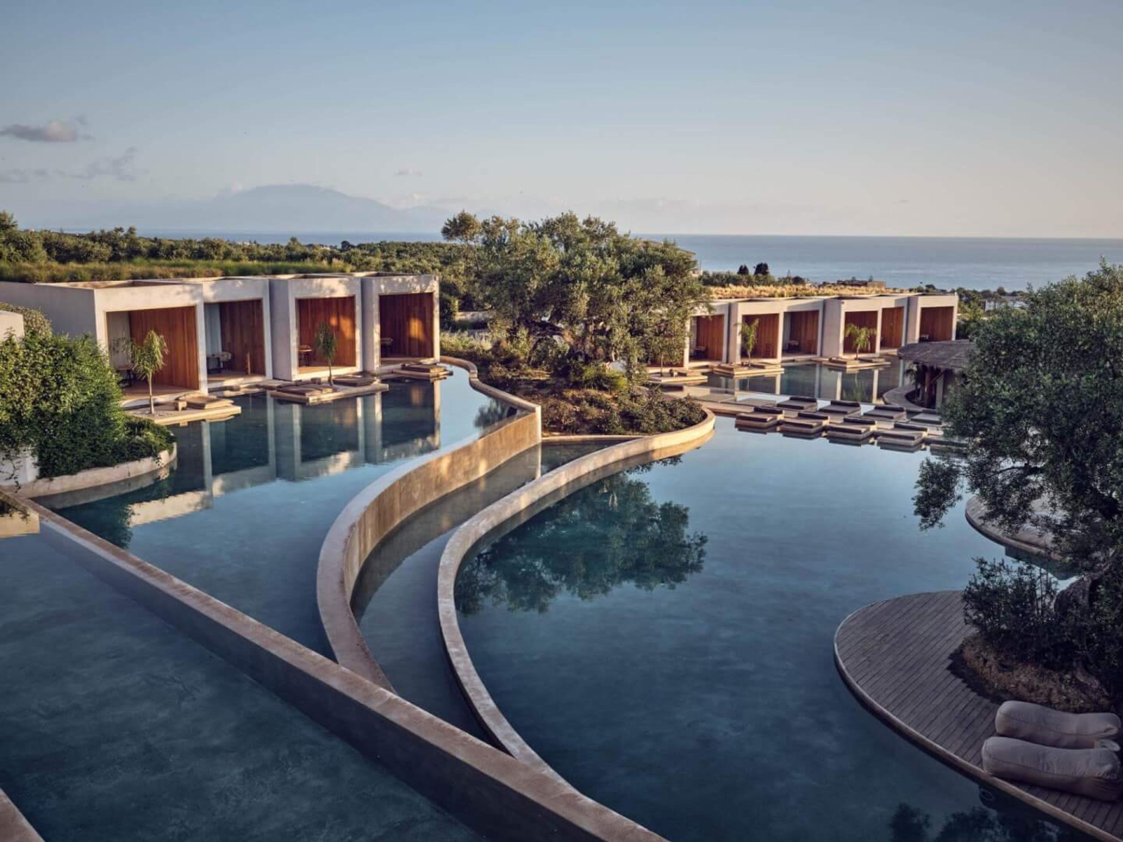 A curving infinity pool lined with guest suites and floating loungers, with the sea and hills beyond under a clear sky.