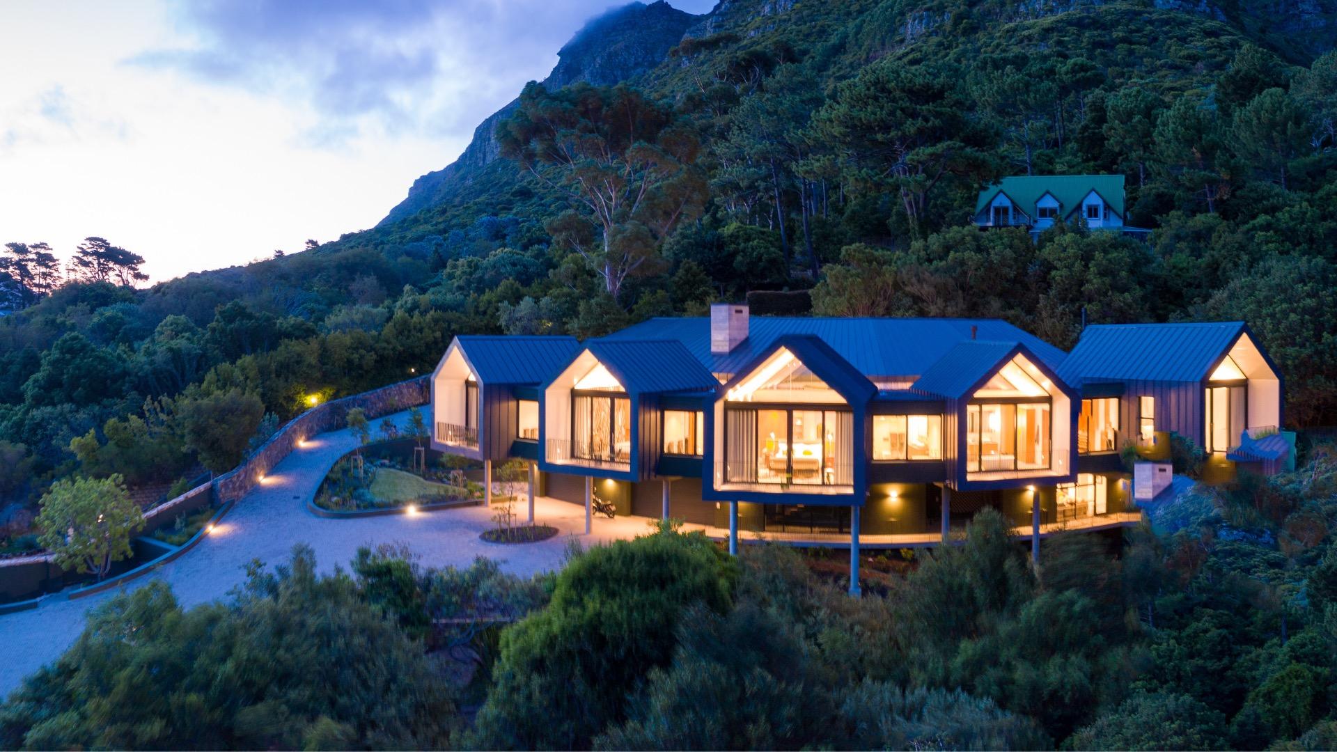 A hillside building with gabled roofs lit from within at dusk, below a forested mountain, with a curved driveway leading to the entrance.