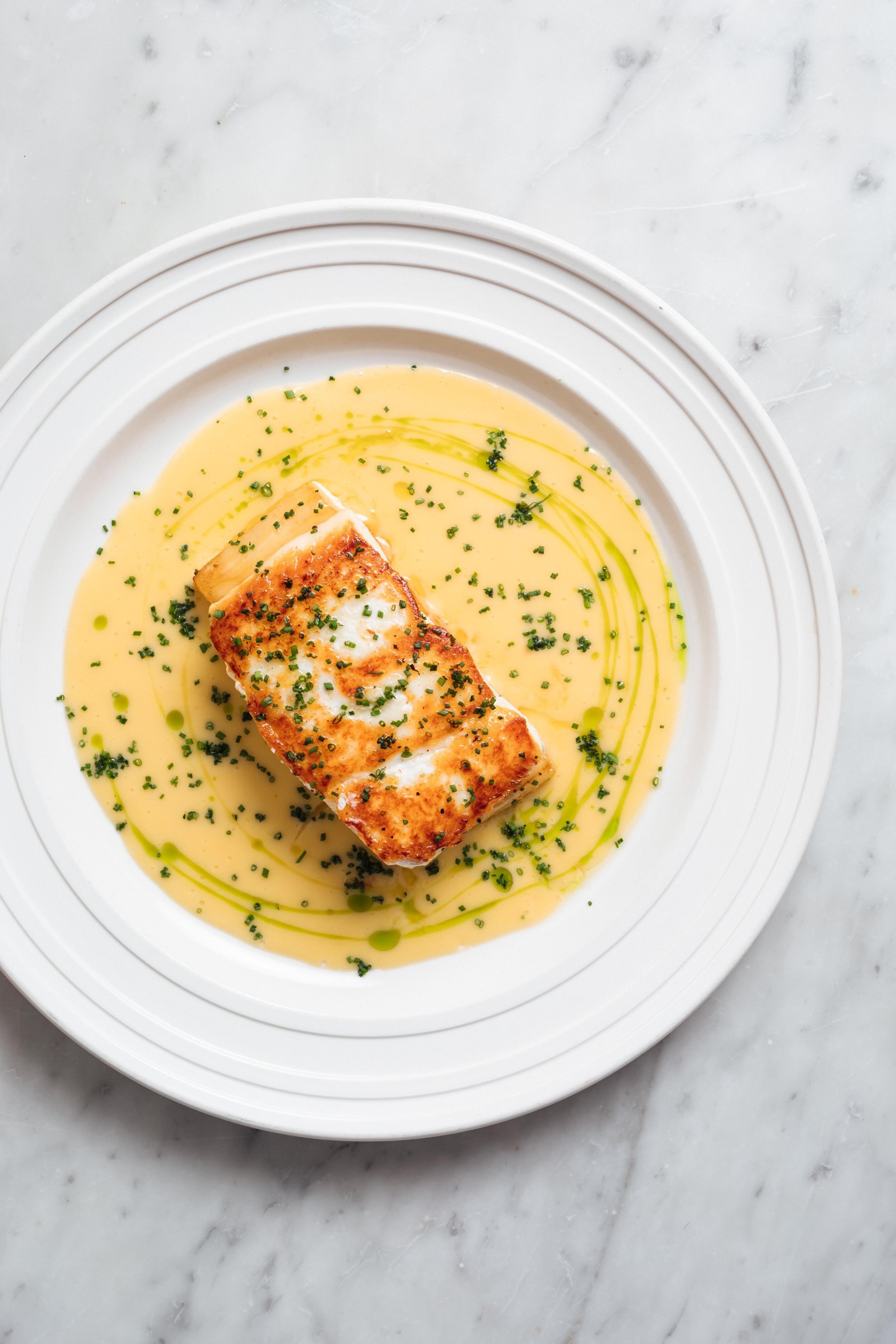A seared fish fillet in a pale butter sauce flecked with chopped herbs, on a white plate.