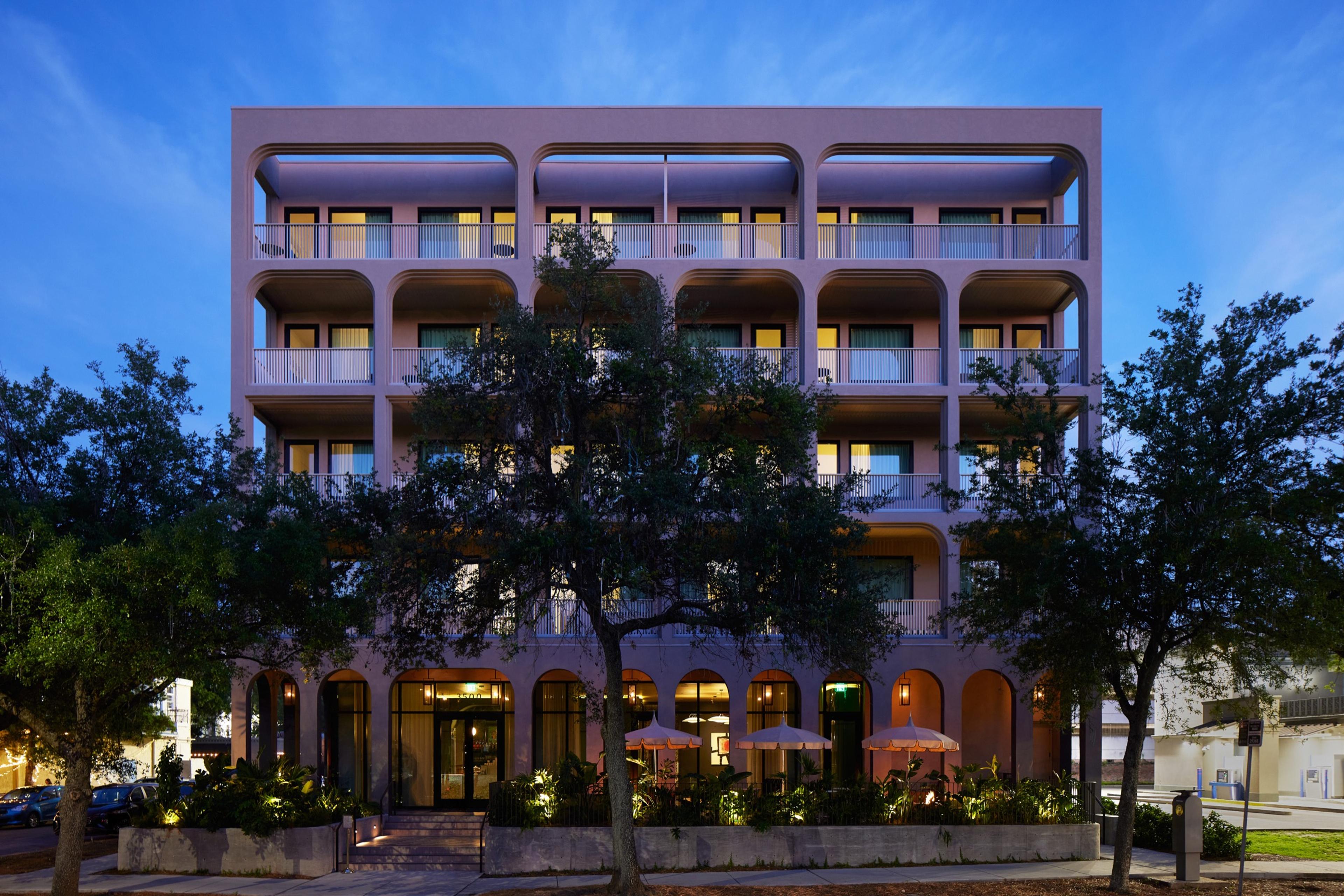 The front of a tall, pale pink building at dusk, with arched balconies on every floor, lit windows, and trees in front of a ground-floor terrace.