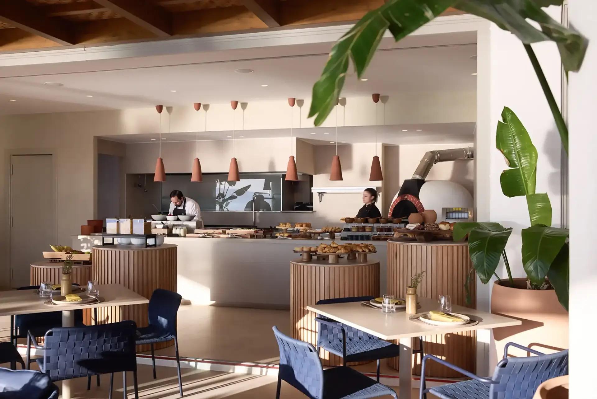 A restaurant's open kitchen with two chefs at the counter, pastries and bread laid out under a pizza oven, tables set in the foreground.