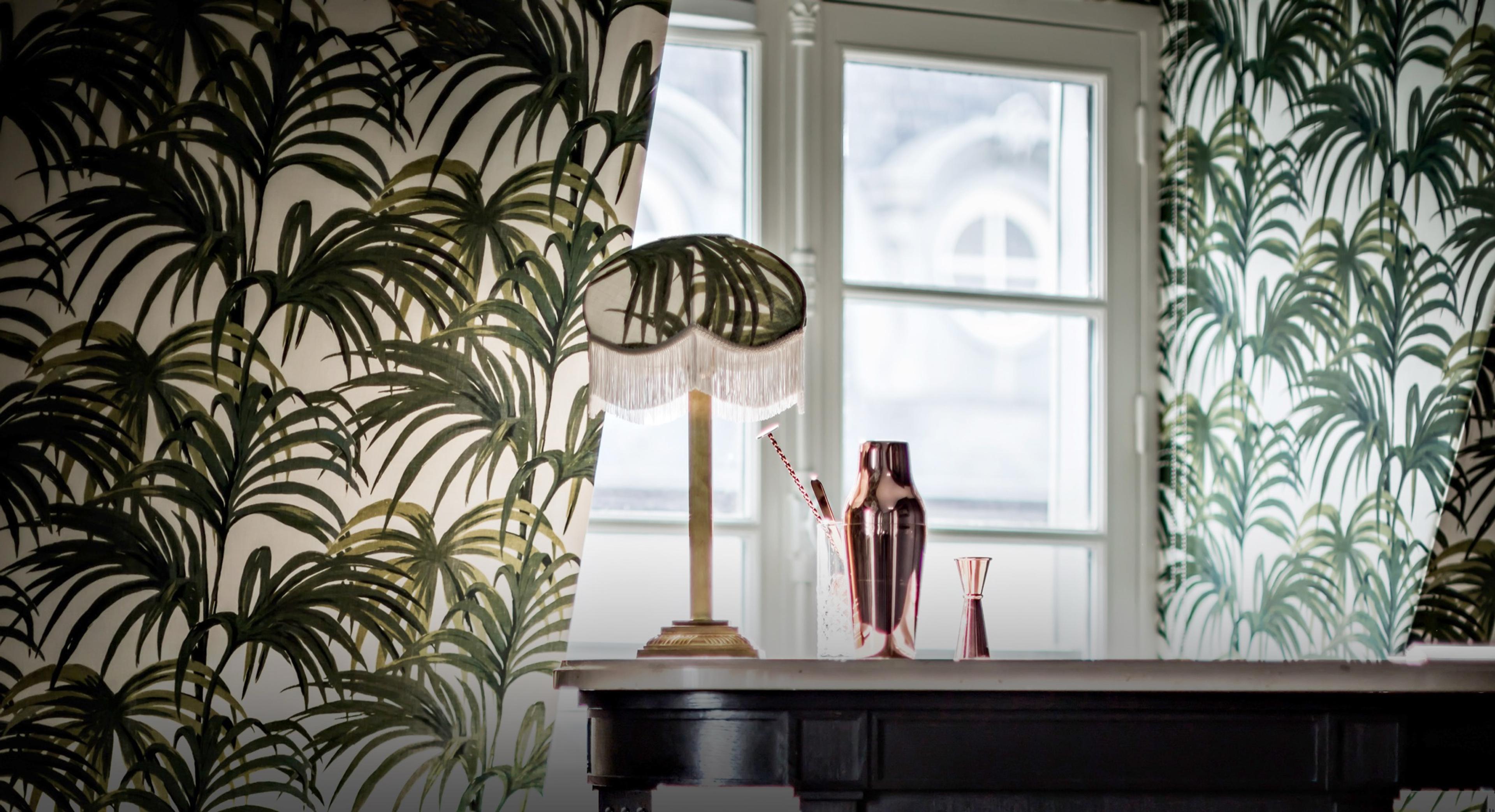 A fringed lamp, a copper cocktail shaker and a jigger stand on a dark console below a window, framed by palm-leaf wallpaper on a curved wall.