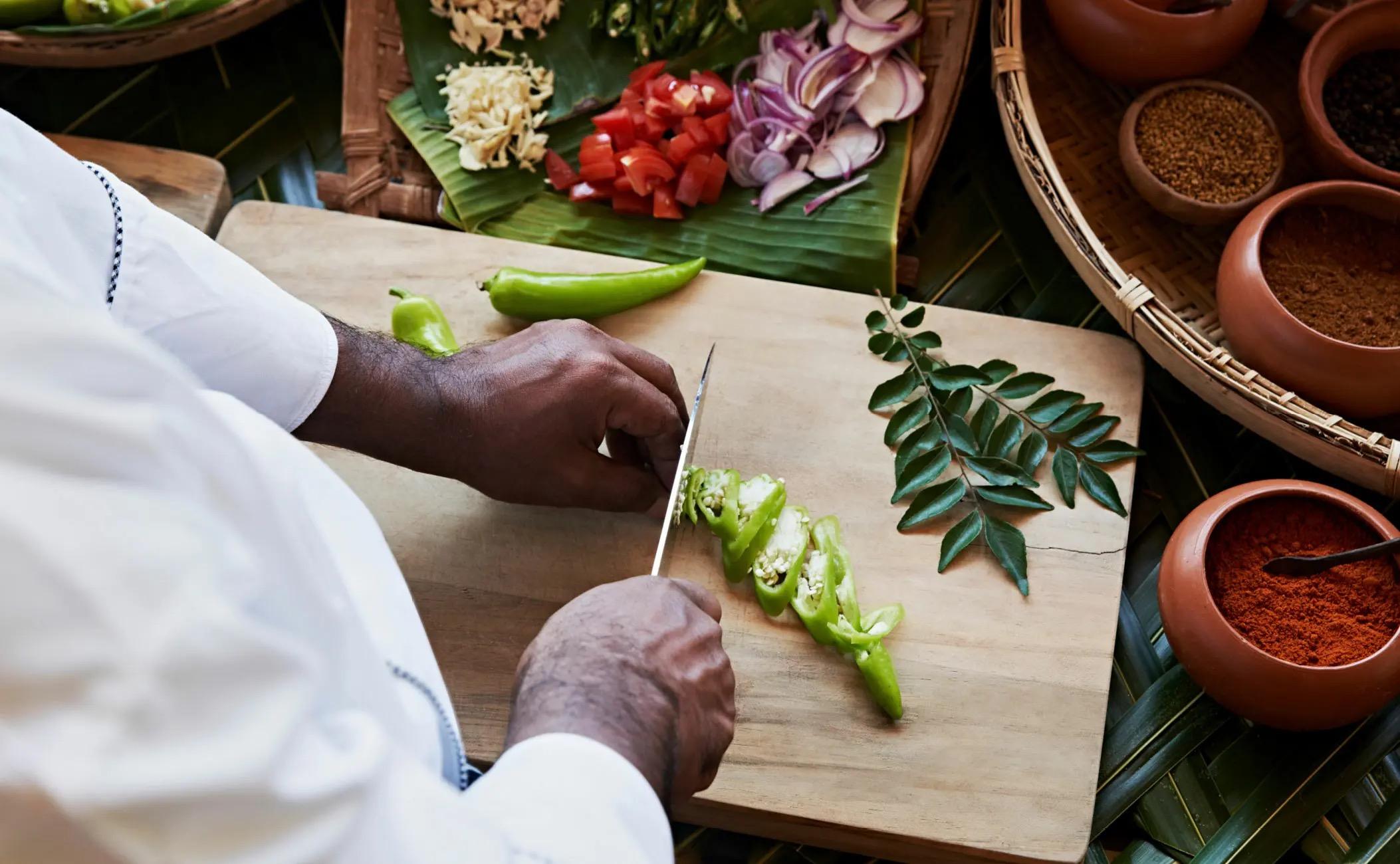 A pair of hands slicing green chillies on a wooden board, with chopped tomato, onion and curry leaves around it.