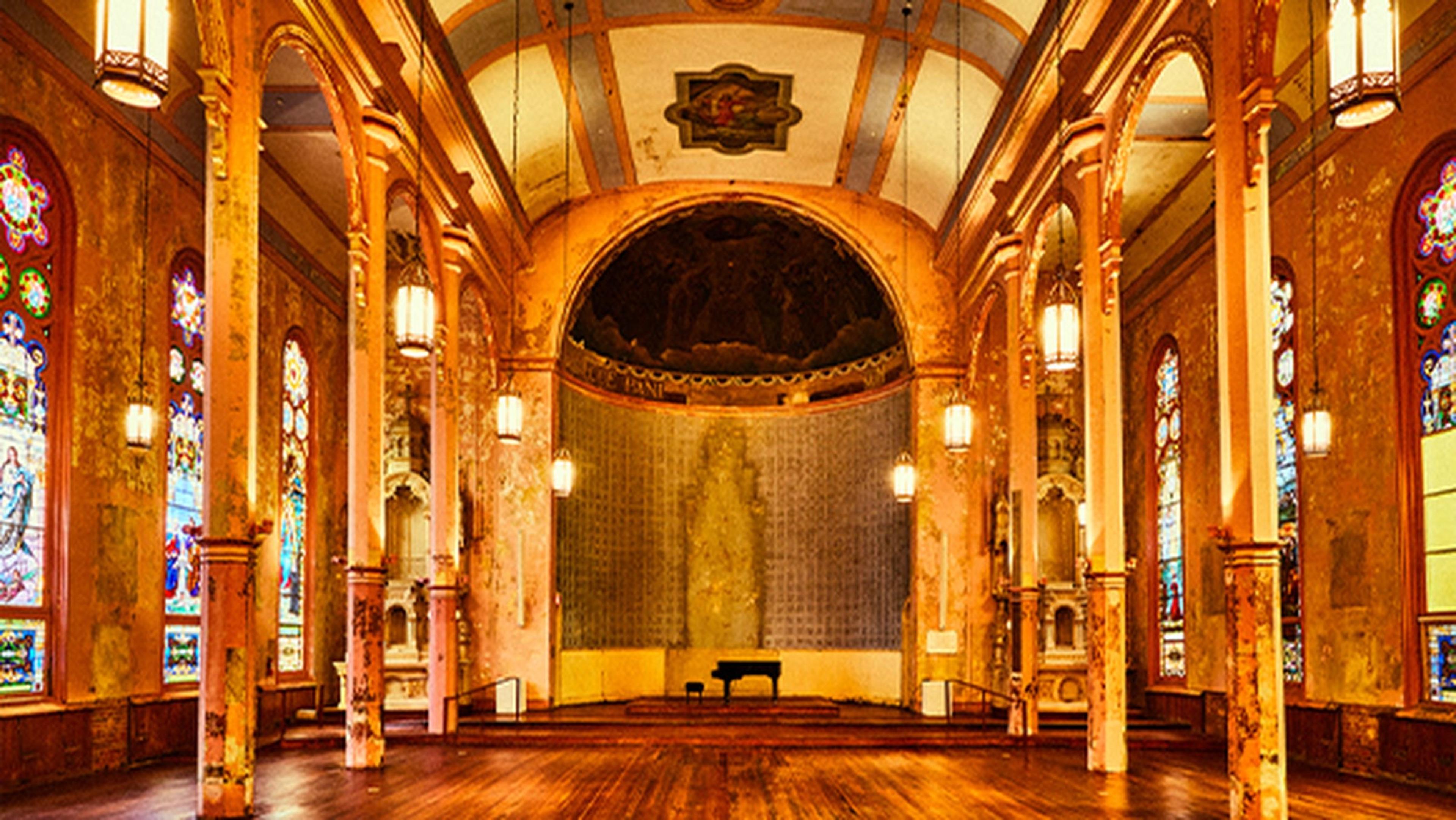 A disused chapel hall with stained glass windows, peeling gilt columns, hanging lanterns, and a piano at the far end beneath a painted dome.