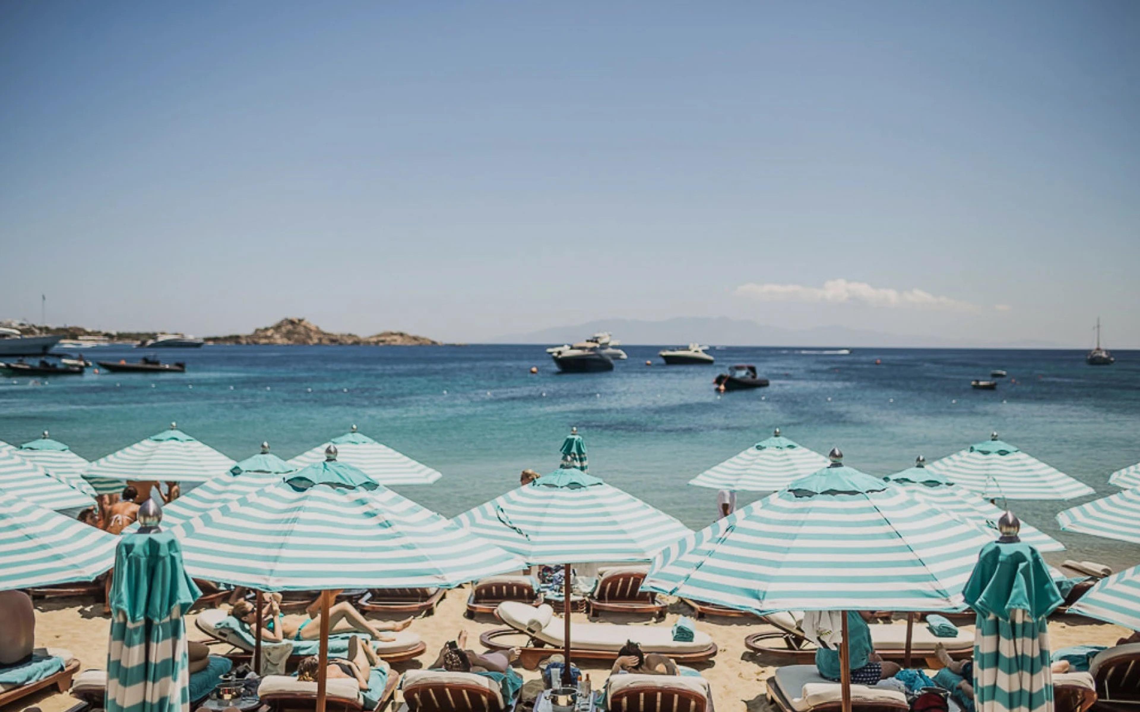 Rows of striped umbrellas and sunbeds along the sand at Nammos on Psarou Beach, boats in the bay