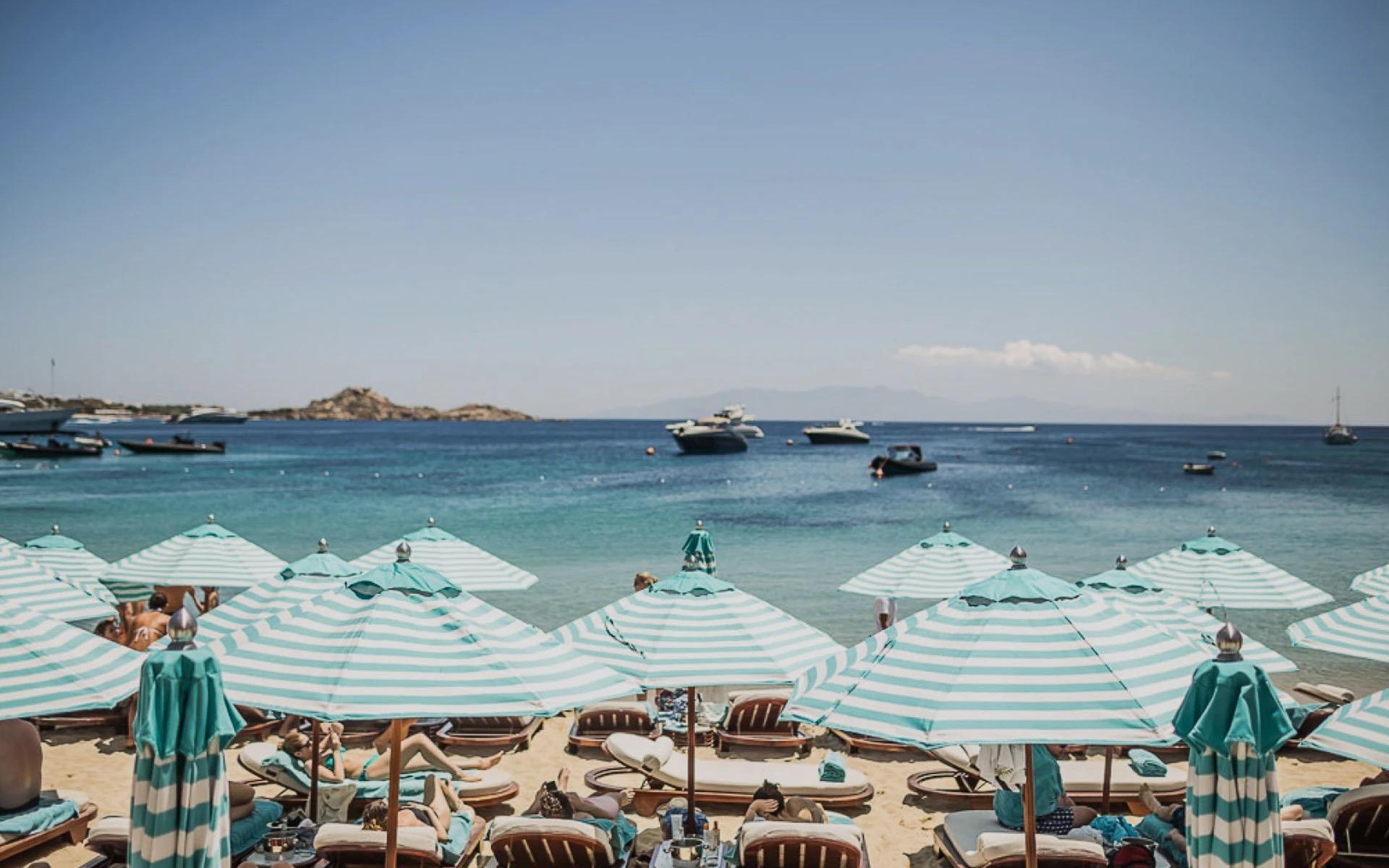 Rows of striped umbrellas and sunbeds along the sand at Nammos on Psarou Beach, boats in the bay