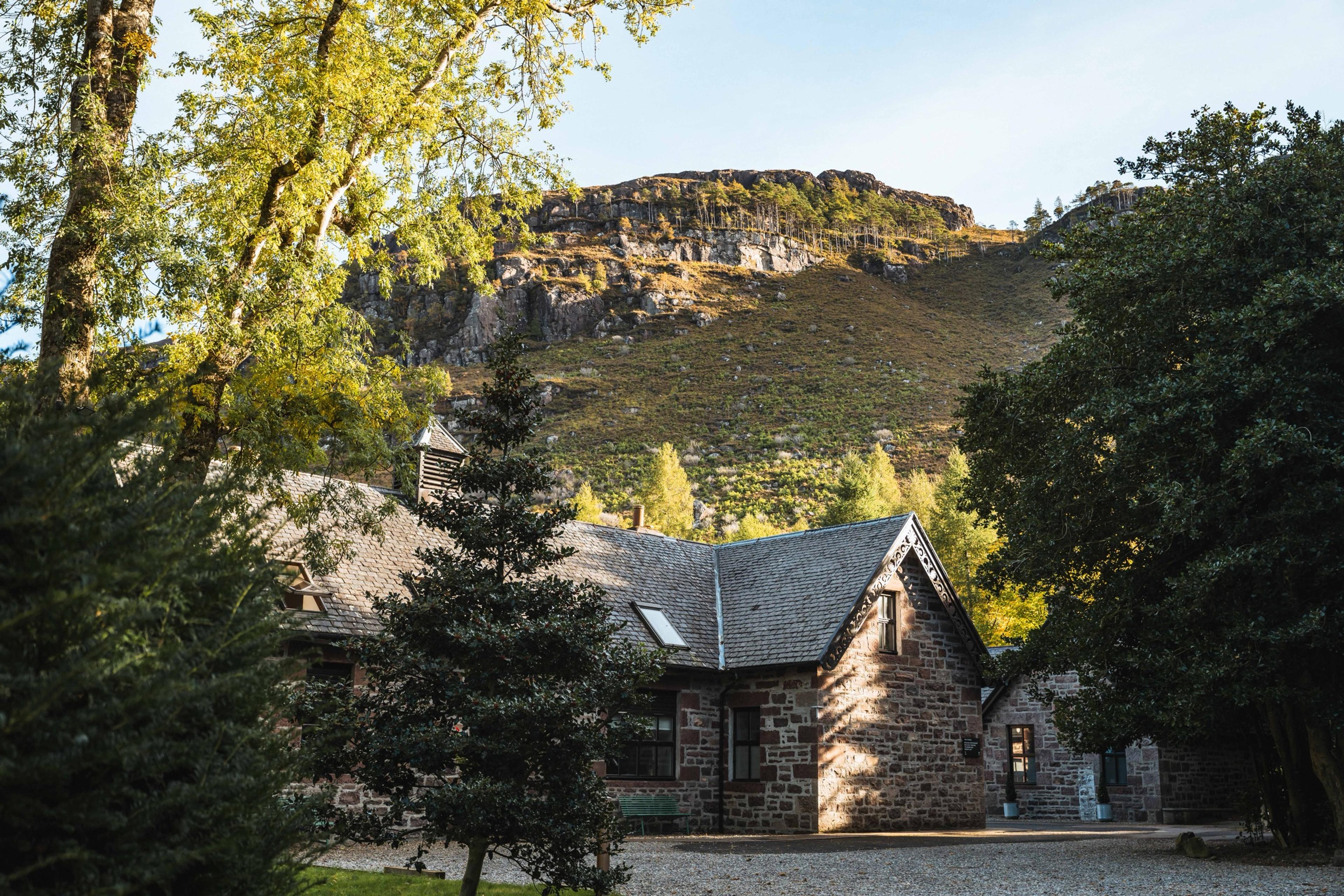 A stone outbuilding with a slate roof among trees, set below a rocky, tree lined hillside.